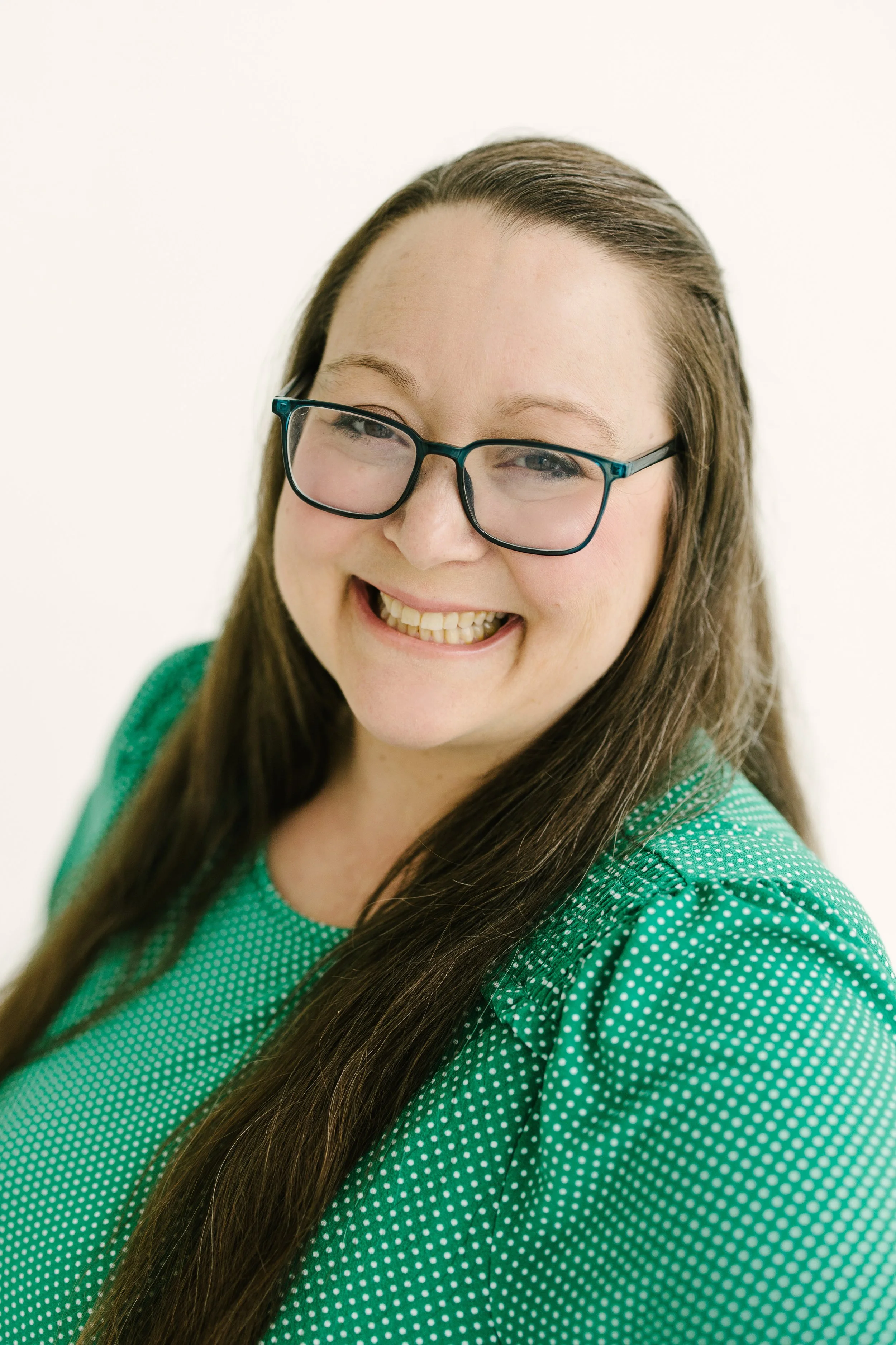 A young woman with long brown hair, wearing glasses and a green polka-dot top, smiling in front of a plain white background.