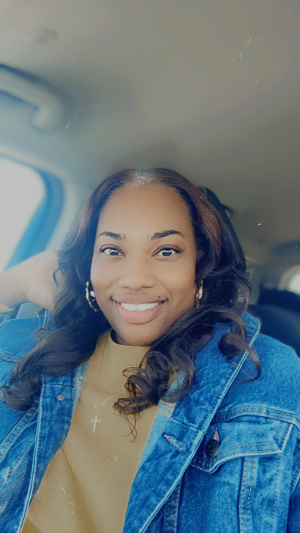 A woman with long, curly hair smiling and sitting inside a vehicle, wearing a denim jacket and a beige top with a cross necklace.