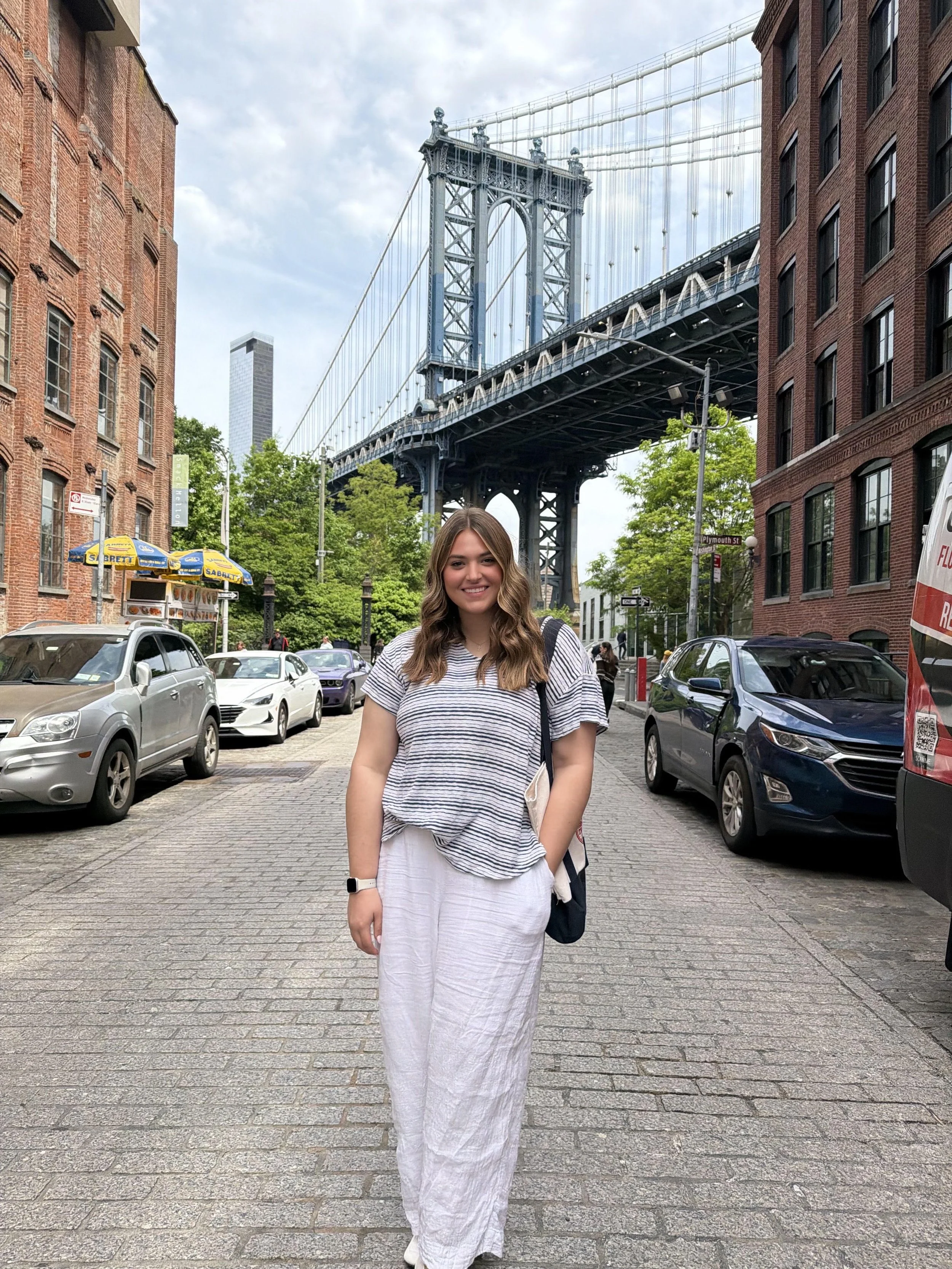 A woman with wavy hair smiling and wearing a striped top and white skirt, standing on a city street with cars, brick buildings, trees, and the Brooklyn Bridge in the background.