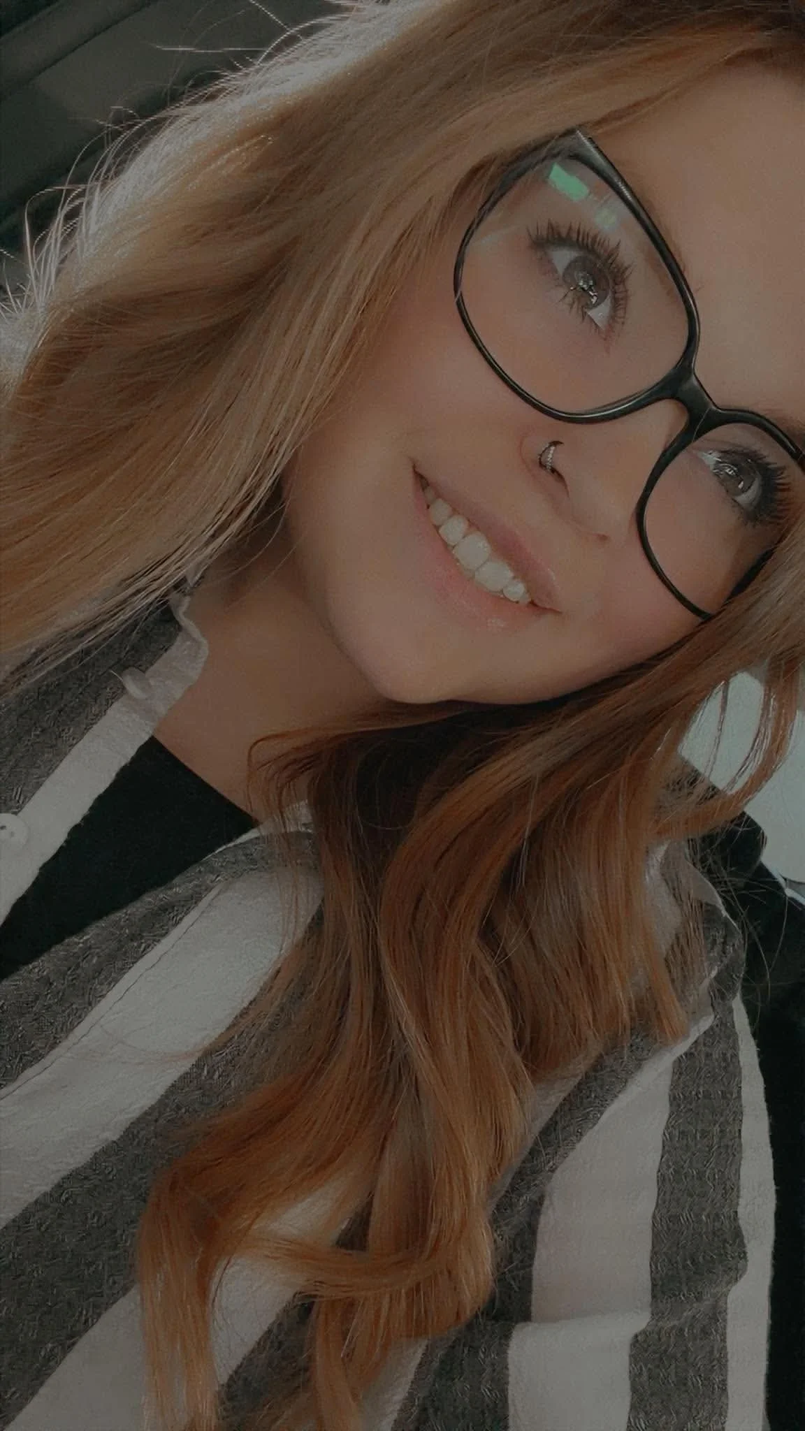 A woman with long, wavy red hair, wearing black-rimmed glasses, a nose ring, and a striped hoodie, smiling and looking to the side.
