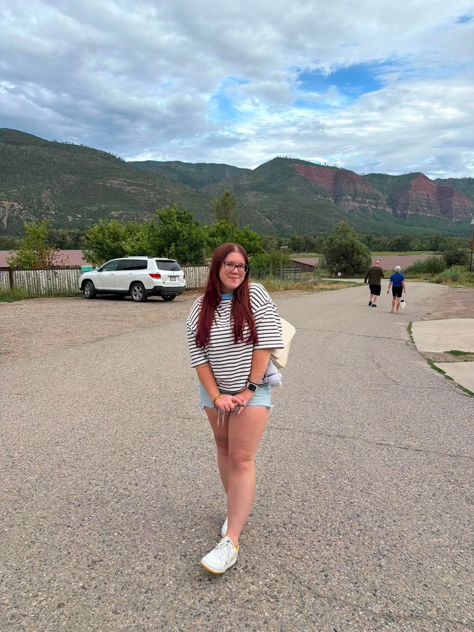A young woman with long red hair, glasses, and a striped shirt standing on a paved area outdoors with mountains in the background. Two older adults are walking away in the distance. A white SUV is parked nearby, and there are trees and small buildings around.