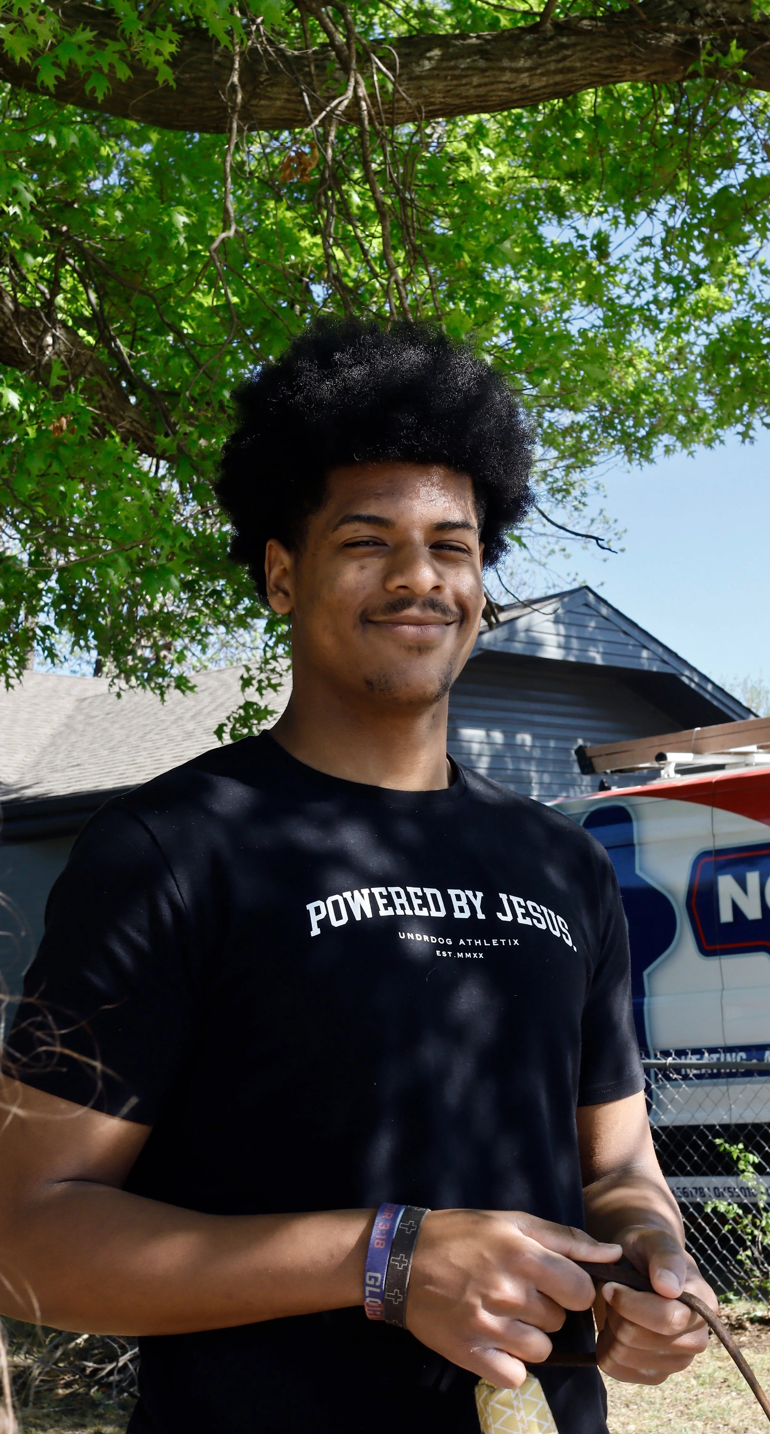 A young man with dark curly hair smiling while standing outdoors under a tree with green leaves. He is wearing a black t-shirt with white text that says 'Powered by Jesus' and some wristbands on his left wrist. He is holding a phone or device in his hands.