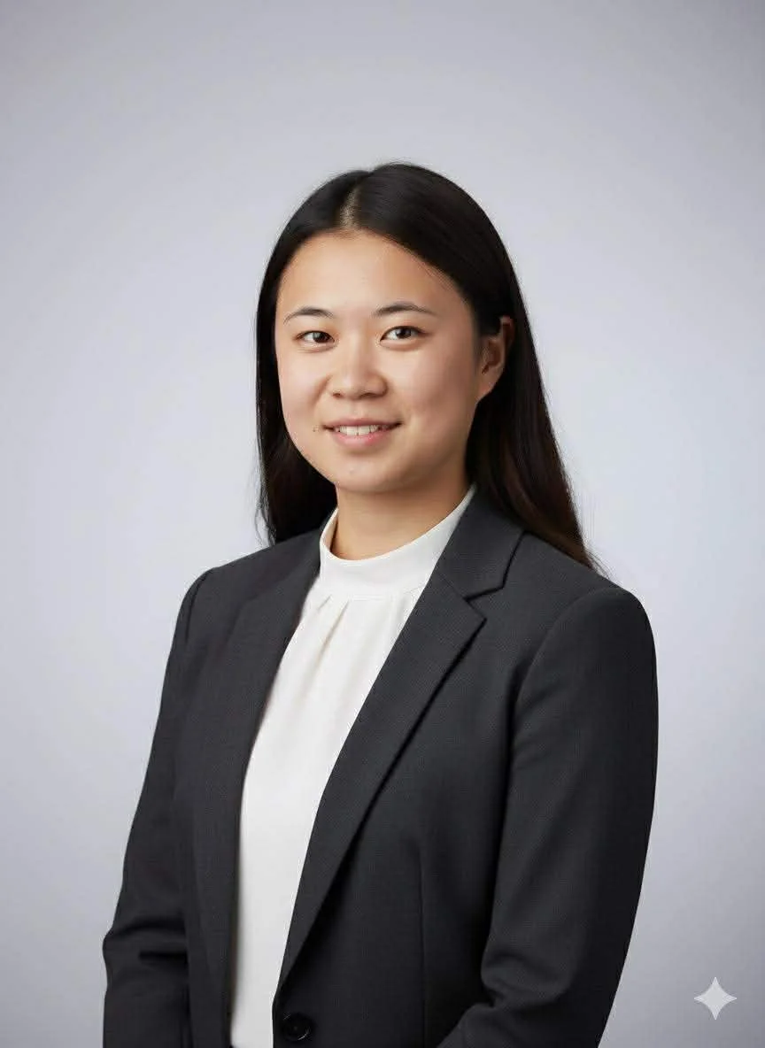 Professional young woman with long dark hair, wearing a black blazer and white blouse, smiling at the camera with a plain light grey background.