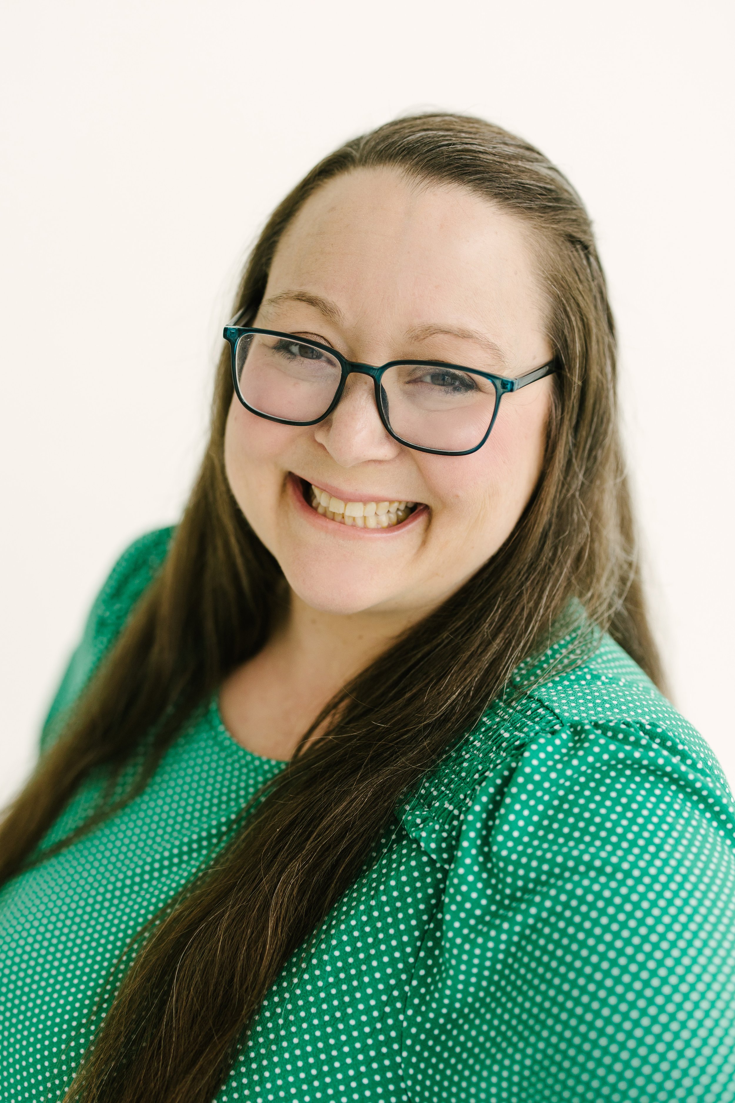 Close-up of a young woman with long brown hair wearing glasses and a green polka-dot shirt, smiling with teeth showing.