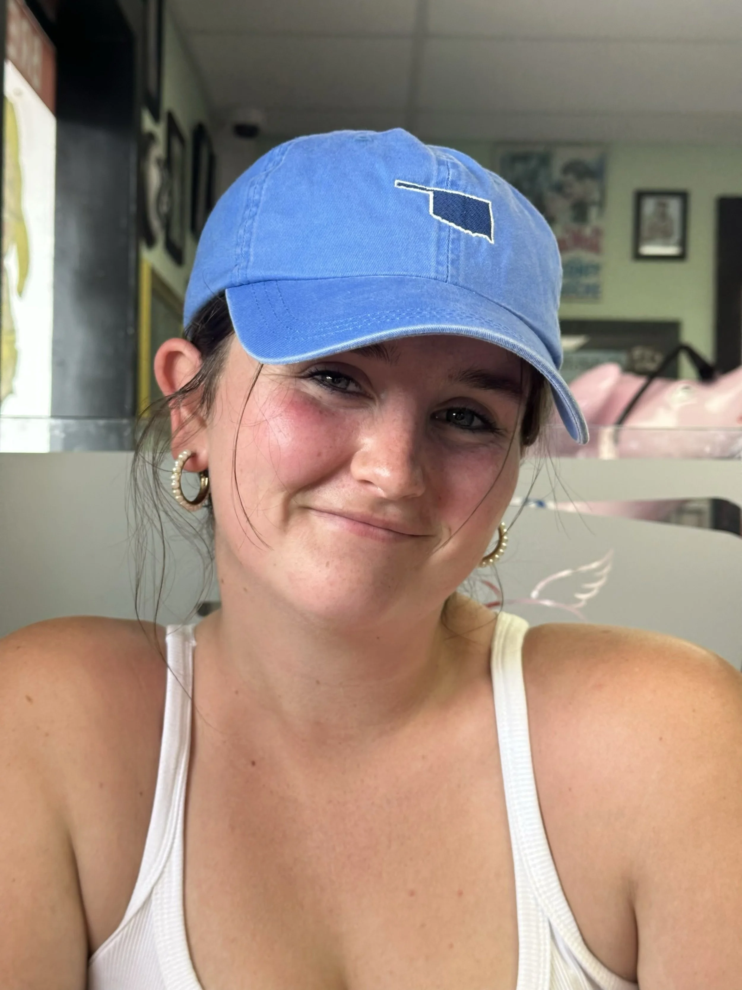 Close-up of a young woman wearing a blue cap with a small patch and a white tank top, smiling, with earrings and freckles visible, in a casual setting.