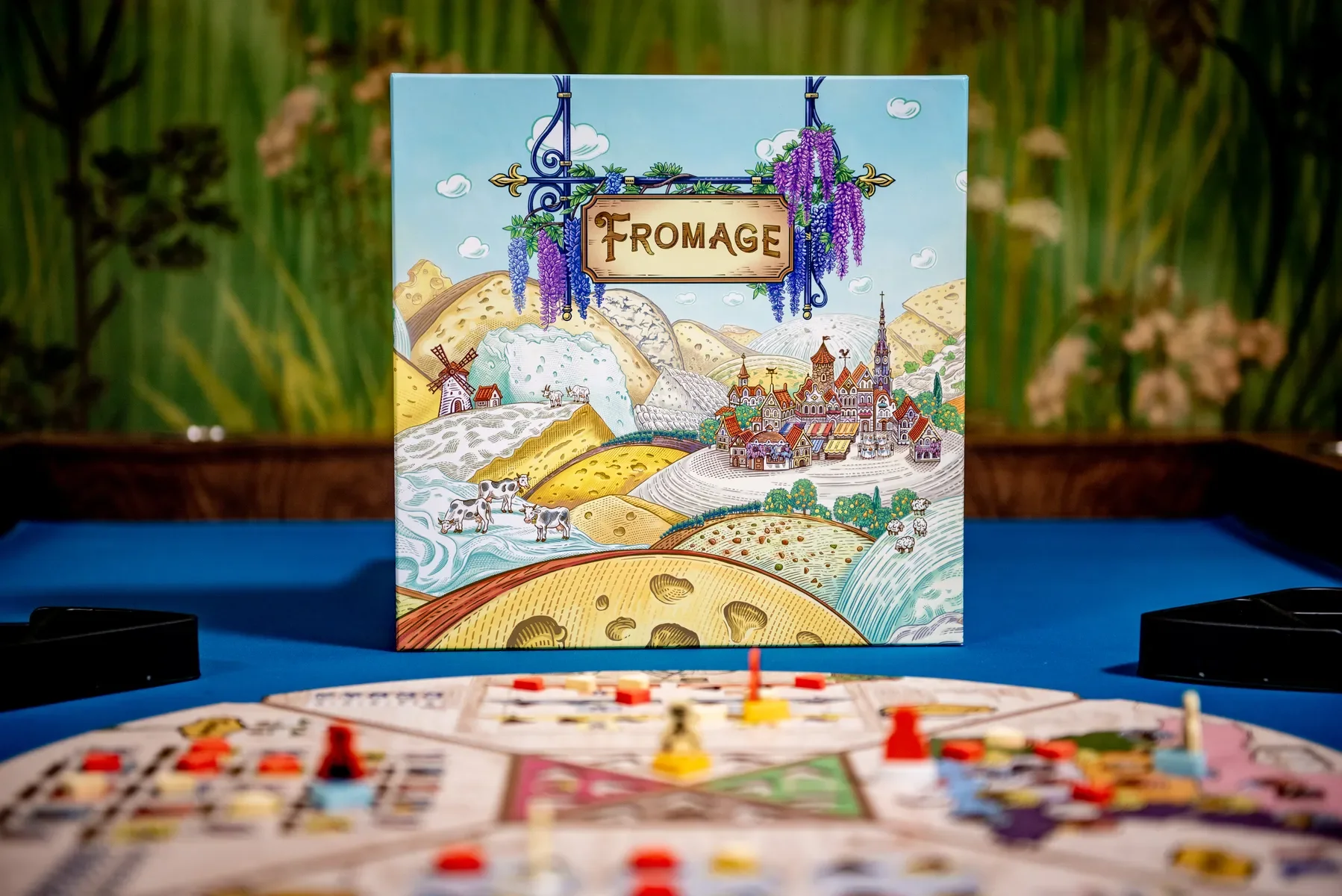 Board game called fromagerie with colorful illustrated cover featuring a farm, cows, a village, and rolling hills, set on a blue table.