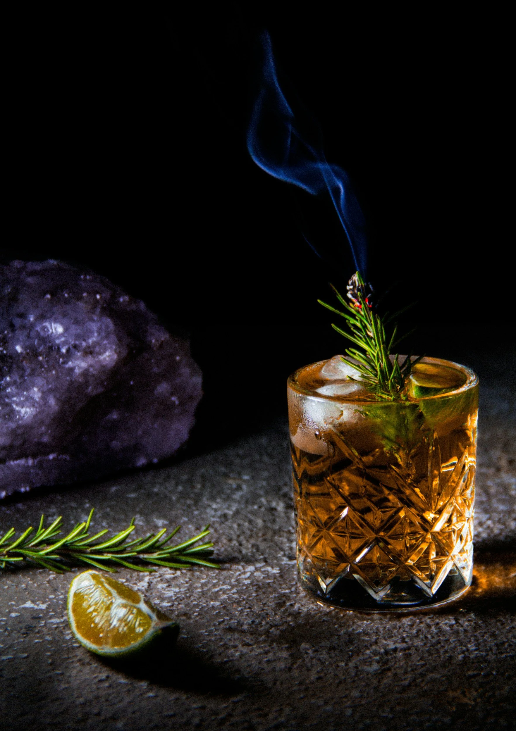 A glass of whiskey with a sprig of rosemary and a slice of lemon on a dark surface, with a rock and lemon slices nearby, and a lighter with smoke rising from it.