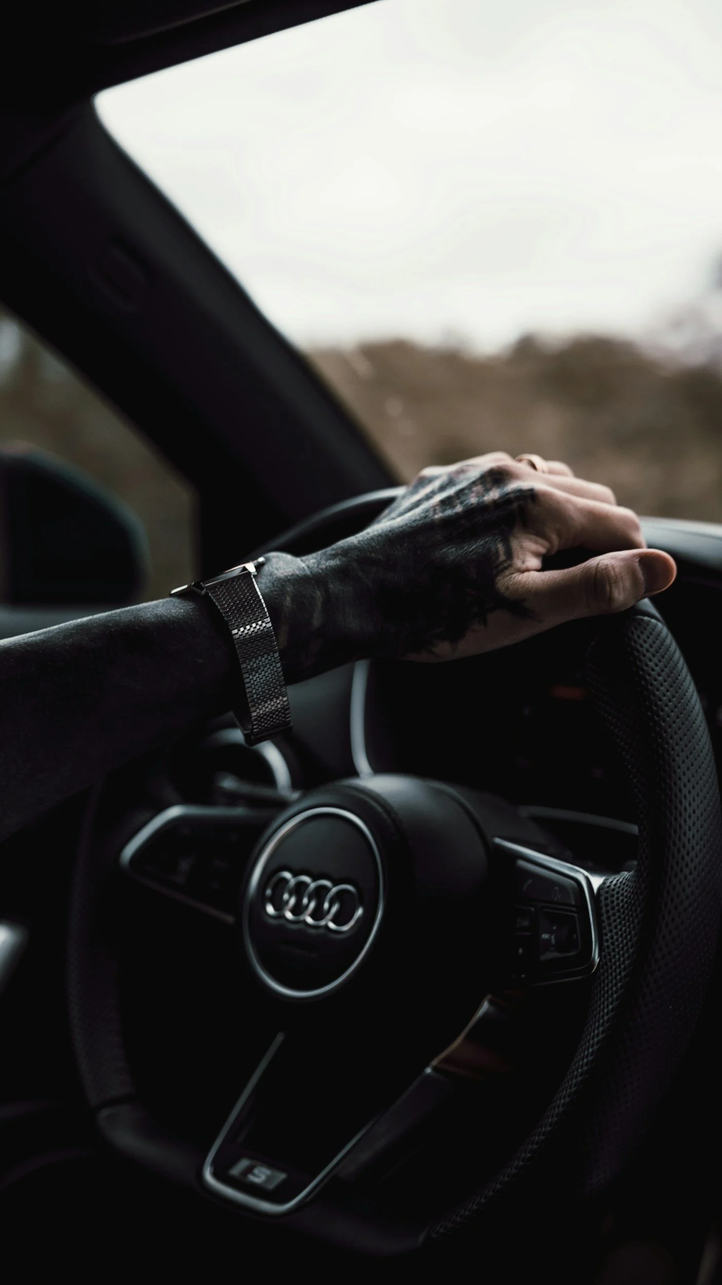 A person's hand with tattoos on the wrist, wearing a watch, resting on the steering wheel of an Audi car, with the dashboard visible. The background outside the car appears to be a blur of nature or landscape.