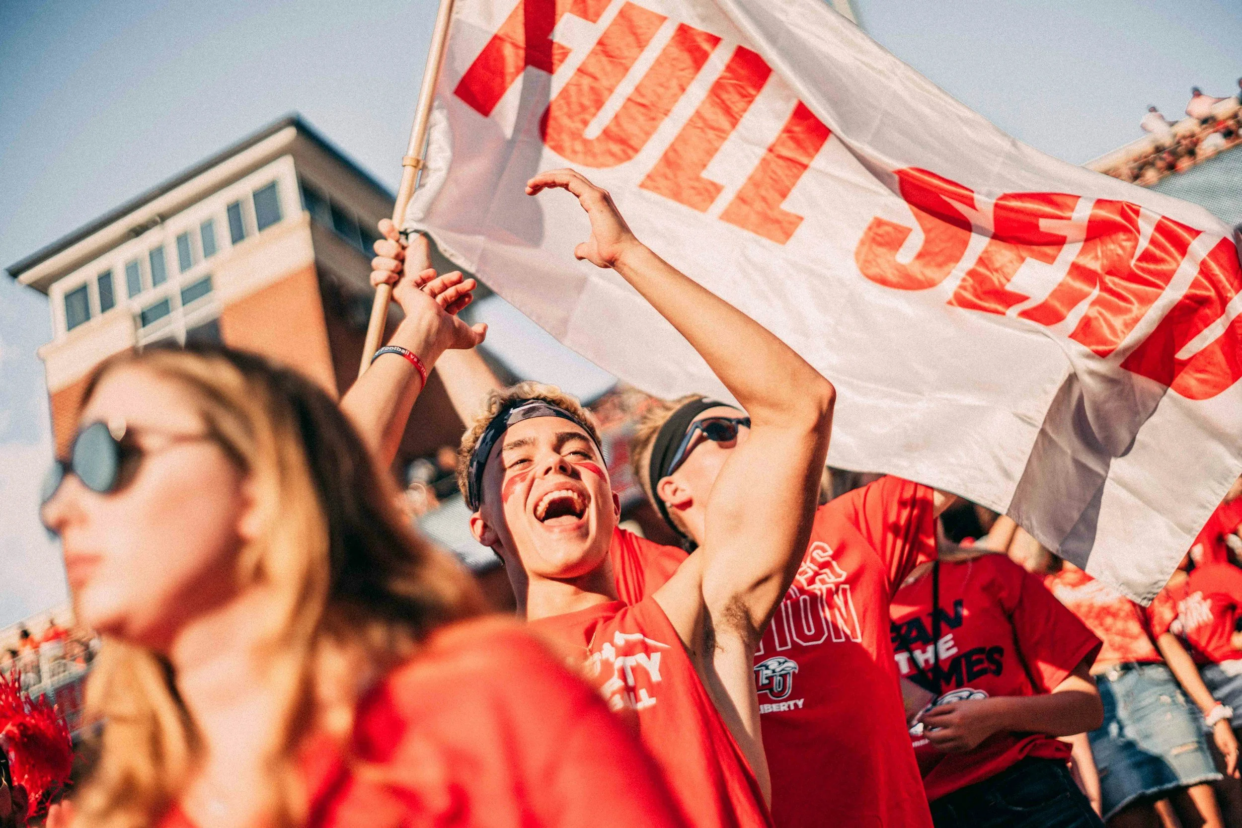 Group of people at a protest or rally, holding a large flag with red lettering, wearing red shirts, and smiling.