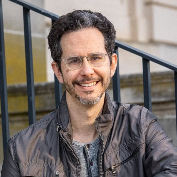 A man with glasses and curly hair smiling outdoors in front of a staircase with a black metal railing.