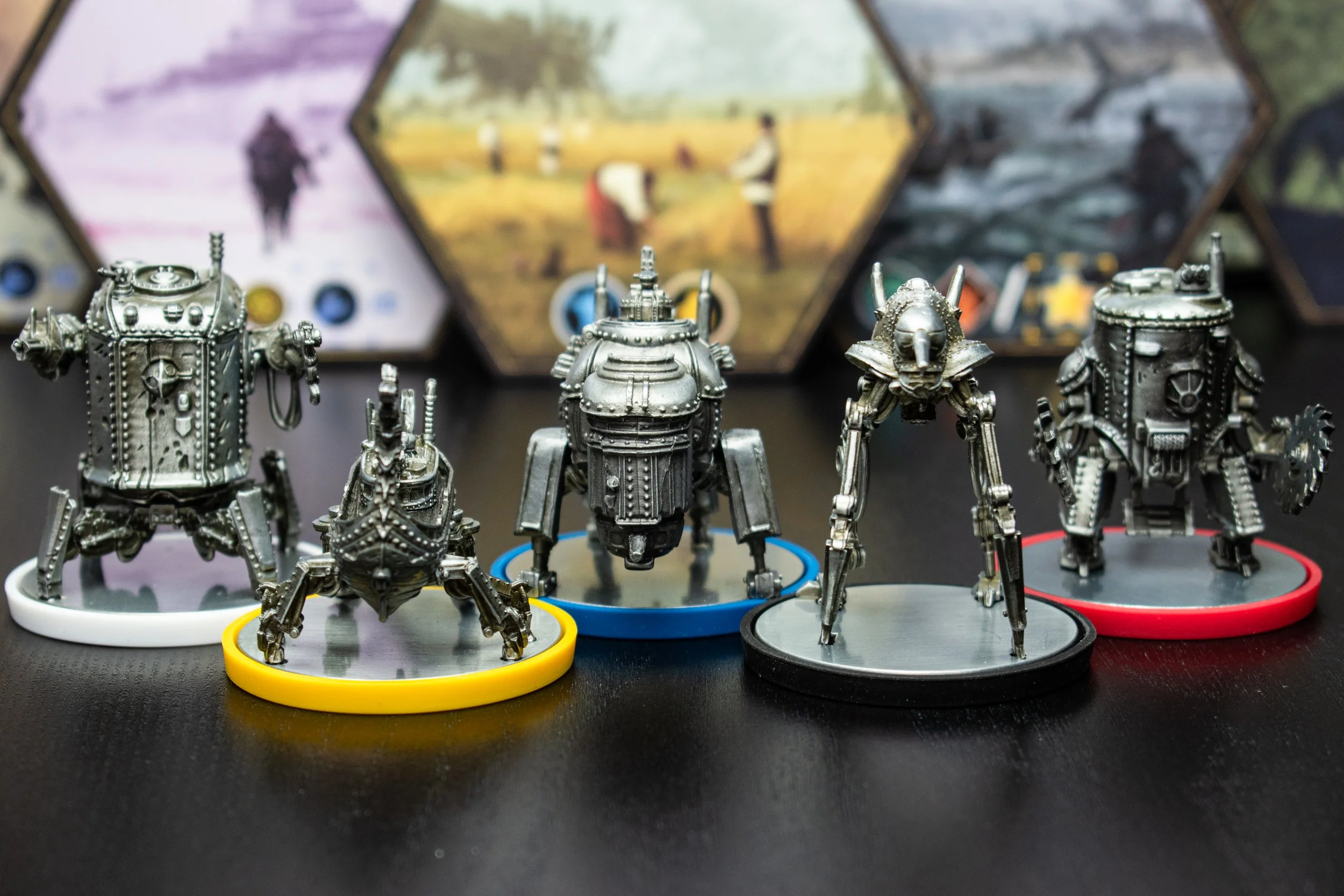A group of metallic robot figurines on small round platforms with colored rims, positioned on a black surface, with blurred images of people and outdoor scenery displayed on hexagonal screens in the background.