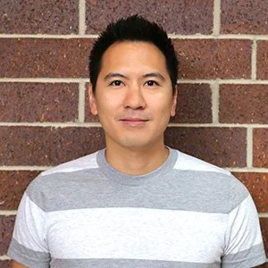 A man standing against a brick wall, wearing a gray and white striped shirt, looking at the camera with a slight smile.