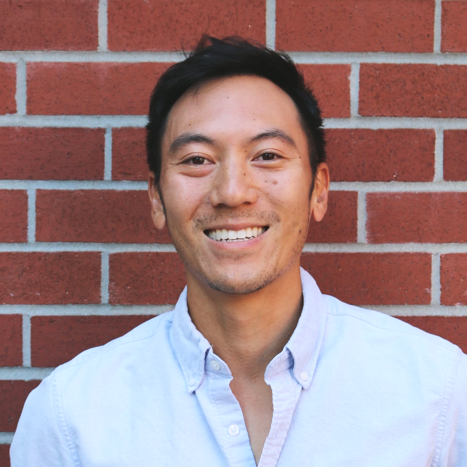 Close-up of a smiling man with dark hair, freckles, and wearing a light blue button-up shirt, standing in front of a red brick wall.