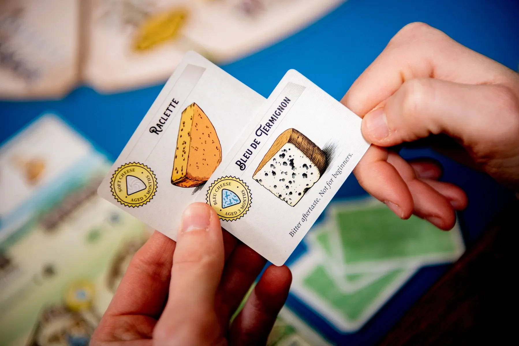 A hand holding cheese tasting cards depicting the types of cheese: Raclette and Bleu de Cevaigne. Each card includes a small illustration of the cheese, its name, and age information.