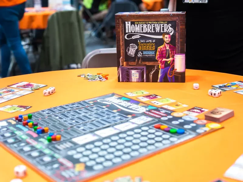A board game called 'Homebrewers' is set up on a yellow table at a gaming event, with game pieces, cards, dice, and game components visible. The game box features an illustration of a man holding a beer mug and a brewing kettle.