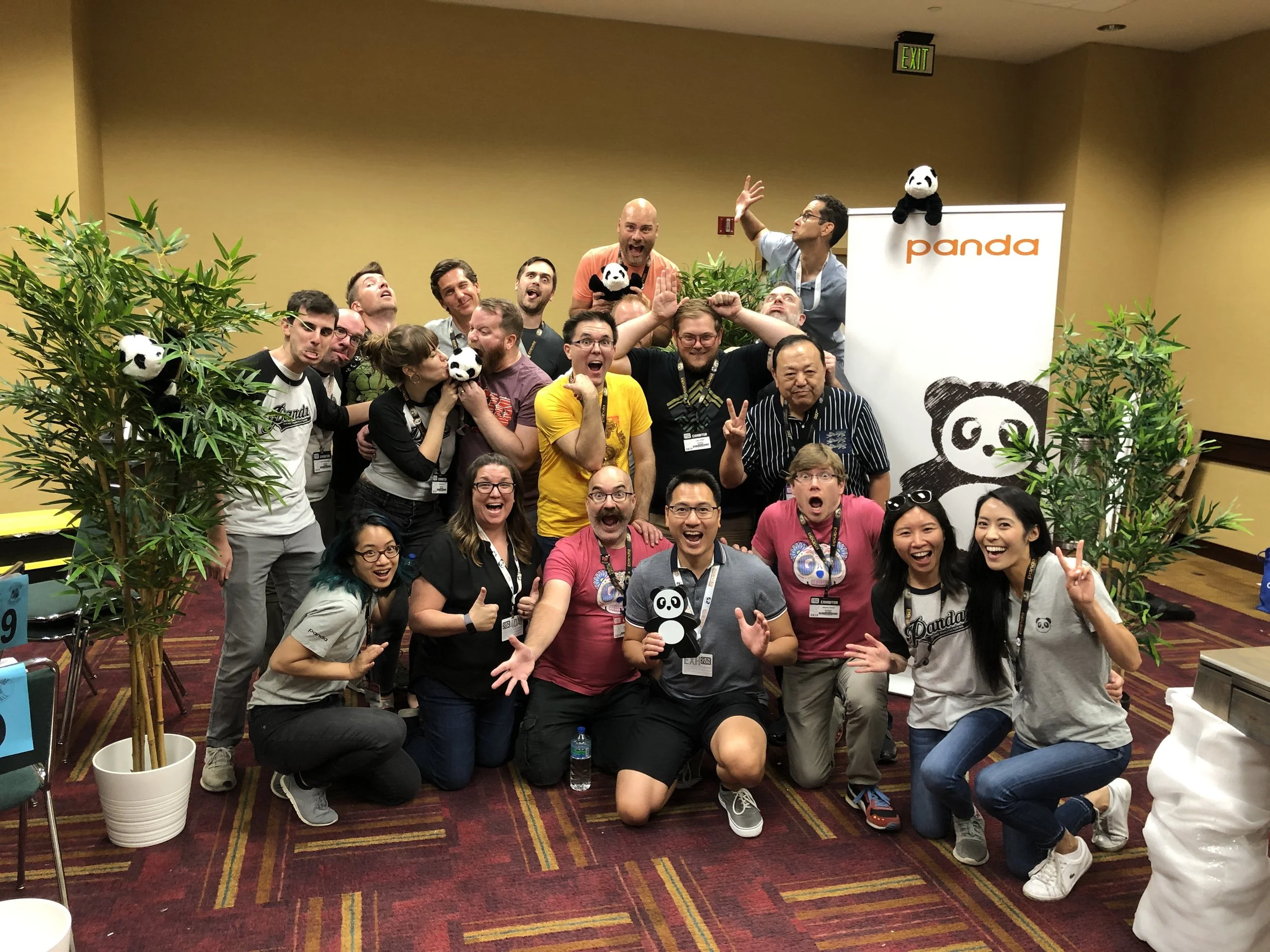 A group of people posing and smiling at an indoor event with a panda theme. The group is diverse, with various ages and appearances, and some are holding or wearing panda plush toys. There is a Panda banner and plants in the background.