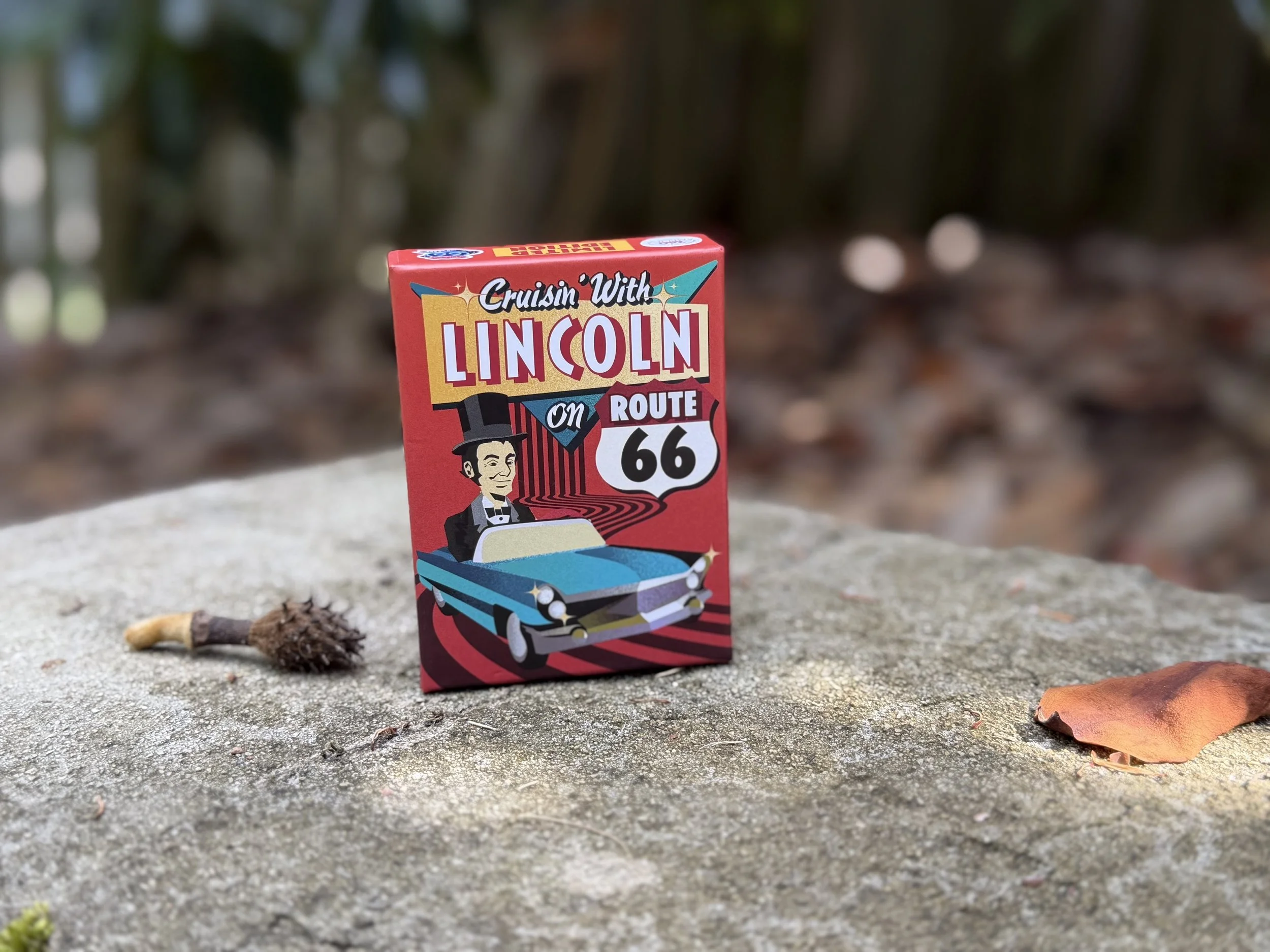 A colorful box of a Crusin' With Lincoln Route 66 game with an illustration of Abraham Lincoln driving a car, placed on a concrete surface outdoors with fallen leaves and blurred greenery in the background.