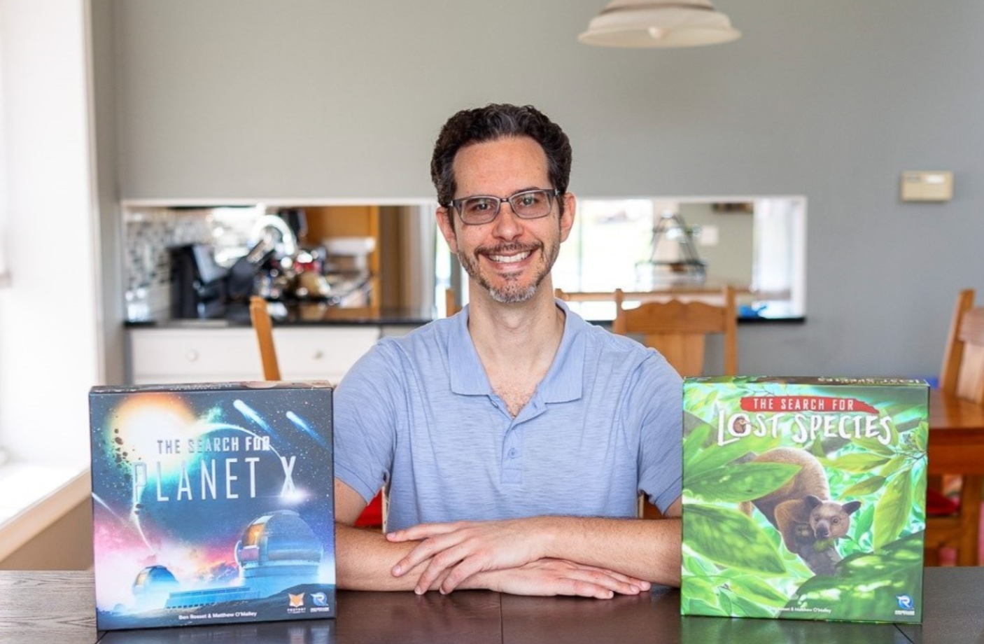 A smiling man with glasses, wearing a blue polo shirt, sitting at a table with two science-themed board games titled "The Search for Planet X" and "The Search for Lost Species."