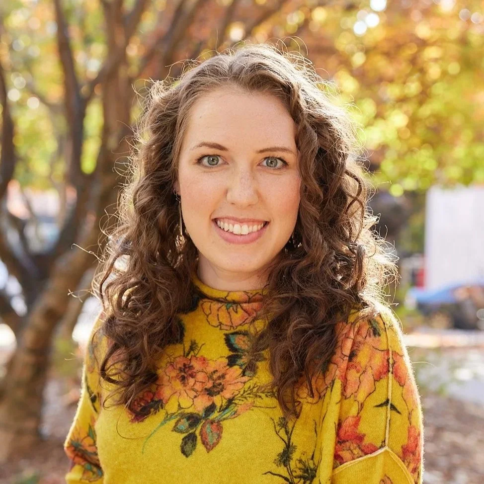 A young woman with curly brown hair and blue eyes smiling outdoors on a sunny day, wearing a yellow floral sweater, with trees and parked cars in the background.