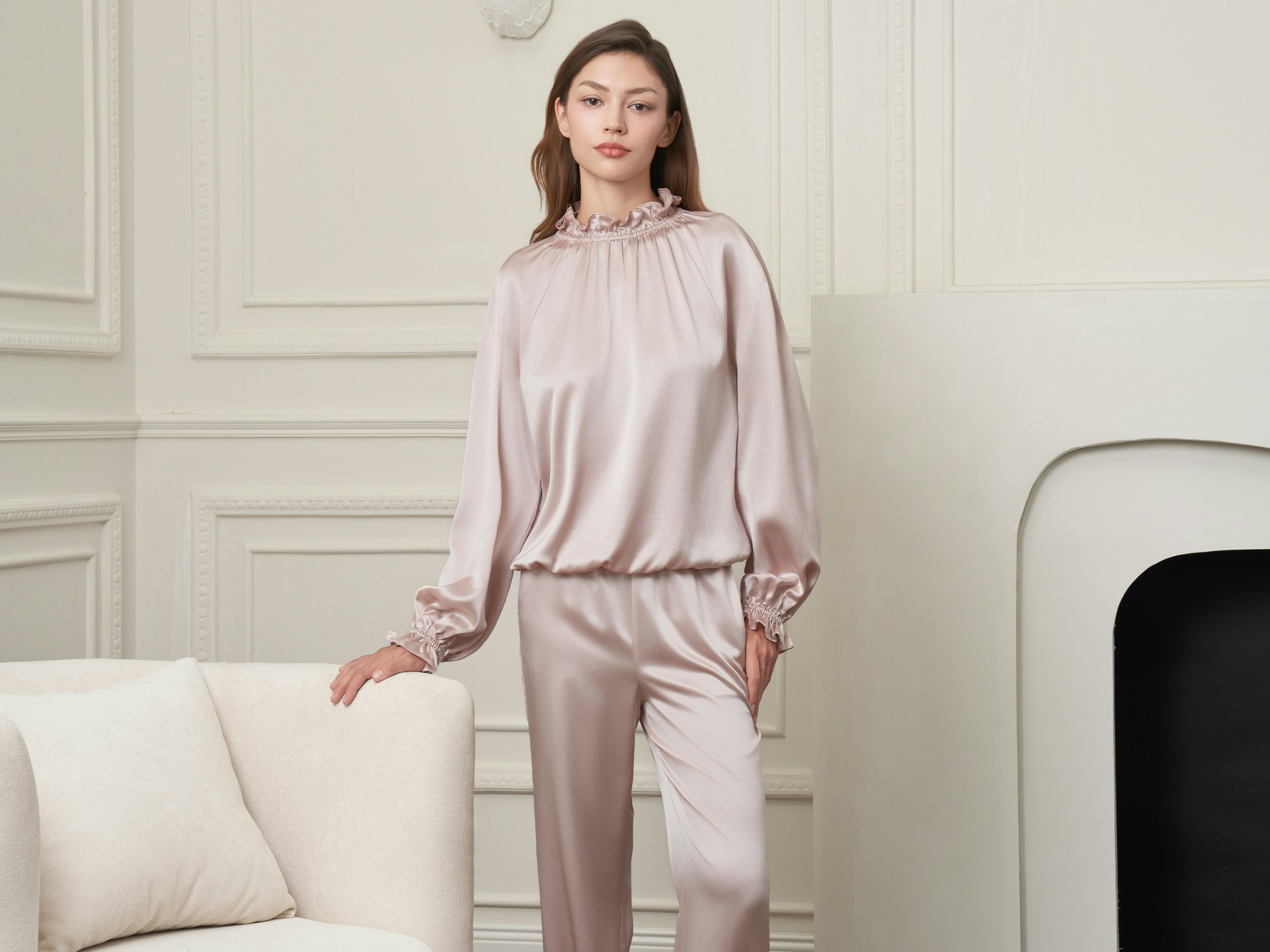 A young woman with brown hair wearing a silky pink satin pajama set, standing in a room with white ornate wall molding, leaning on a white couch with her right hand.