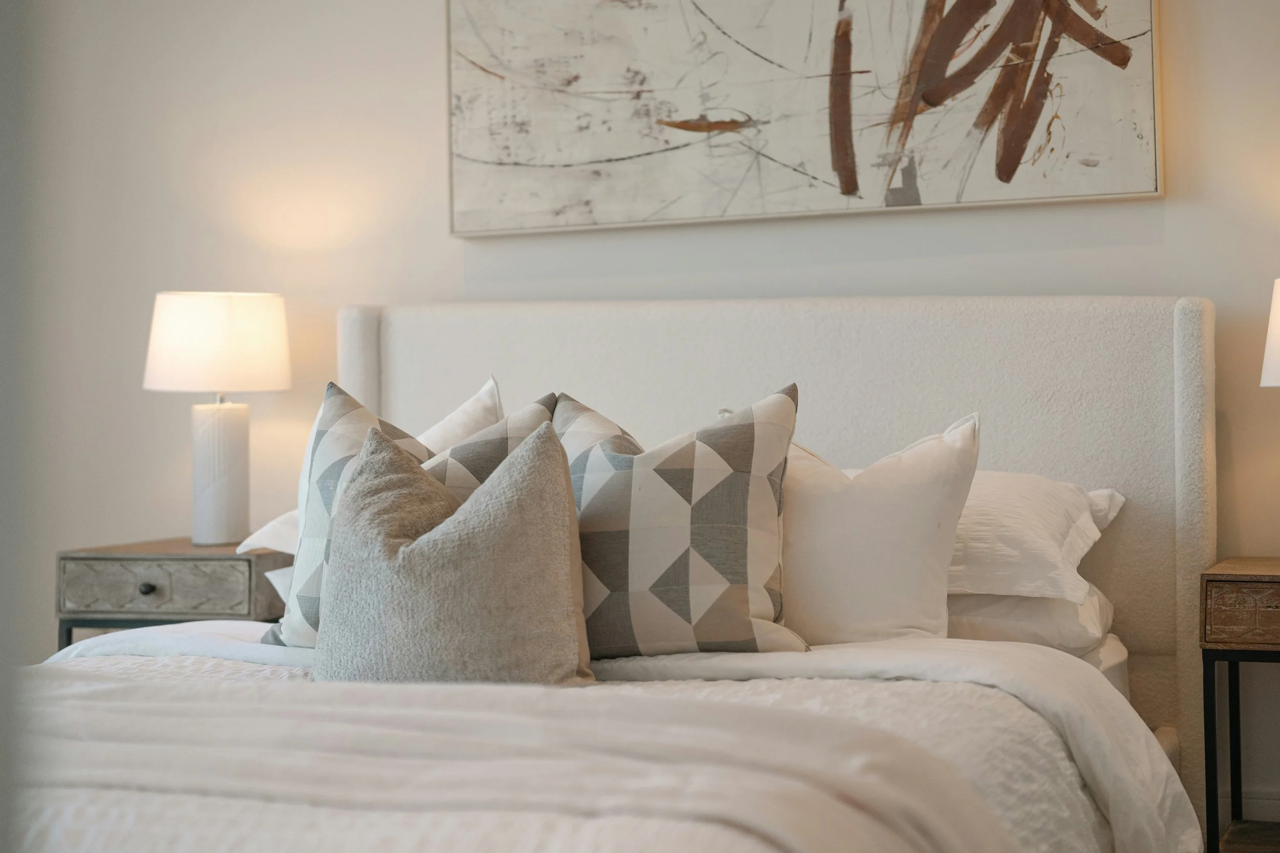 A neatly made bed with numerous pillows and a white headboard in a modern bedroom. A bedside table with a marble top and a lamp are visible on the left. A large abstract painting hangs above the bed, and a nightstand is on the right.
