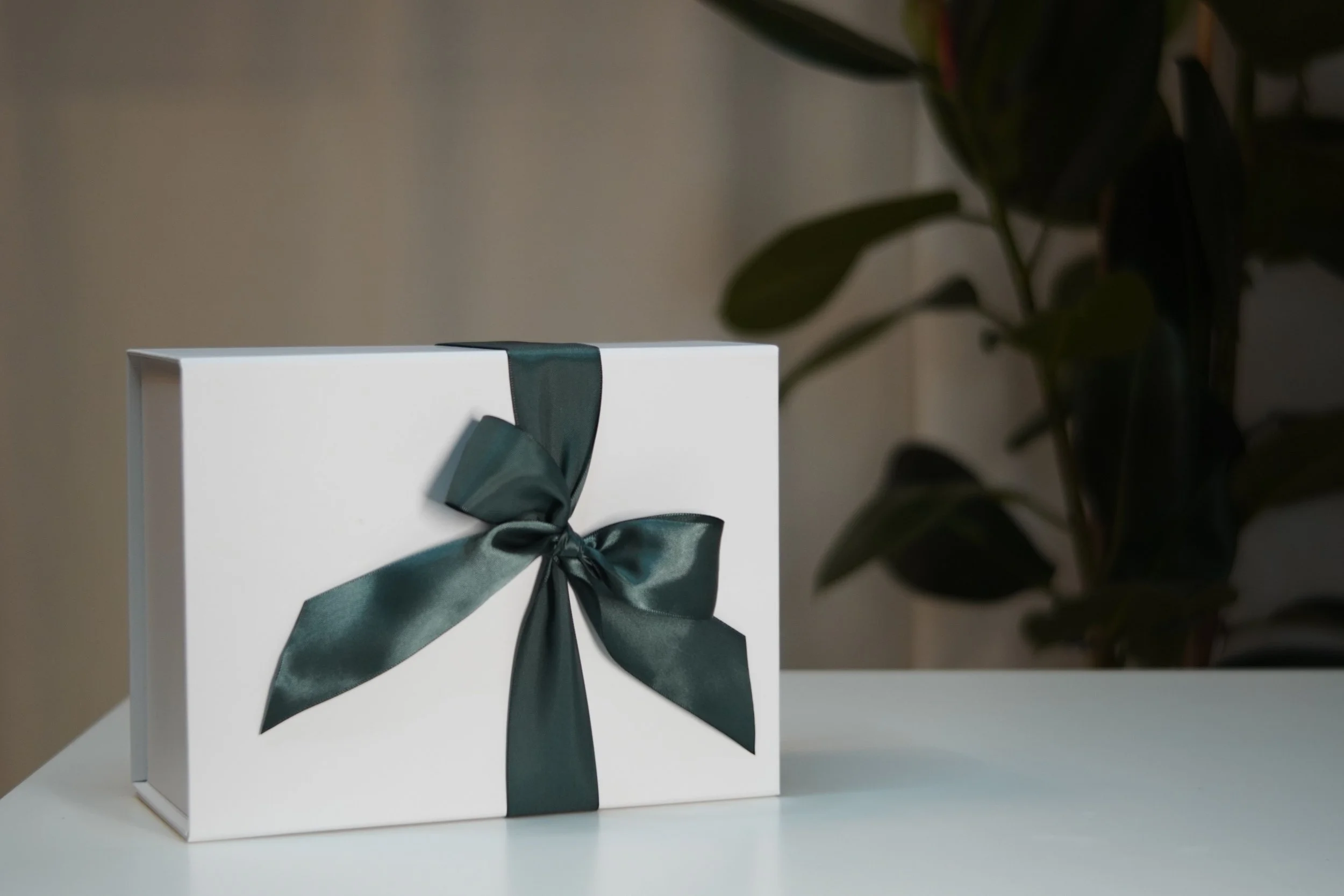 A white gift box with a dark green satin ribbon tied in a bow, placed on a white surface with a blurred background of a plant with dark green leaves.
