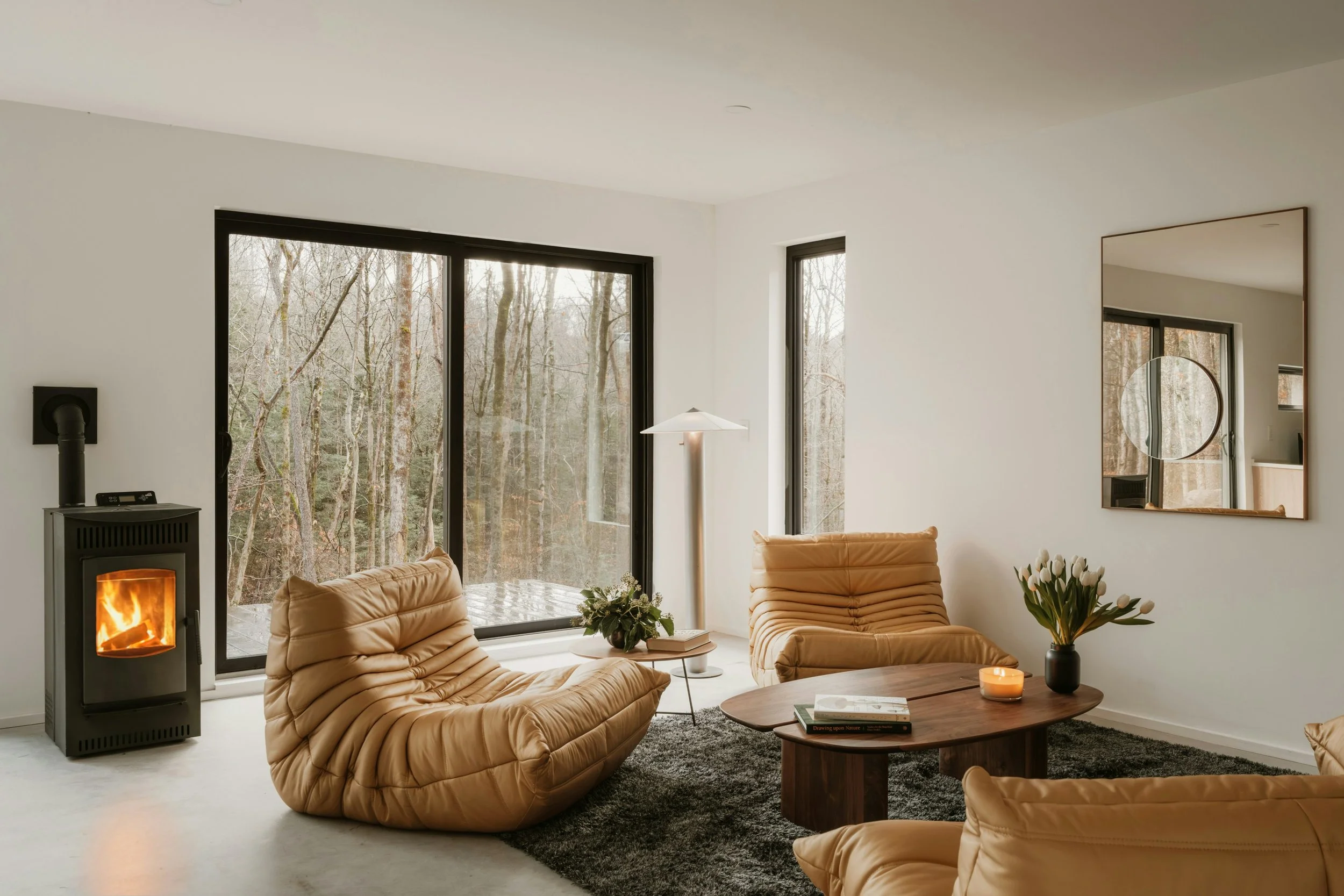 Modern living room with beige leather recliner chairs, a black fireplace, large windows showing a wooded outdoor view, a round wooden coffee table, a tall mirror, a white floor lamp, and decorative flowers in a black vase.