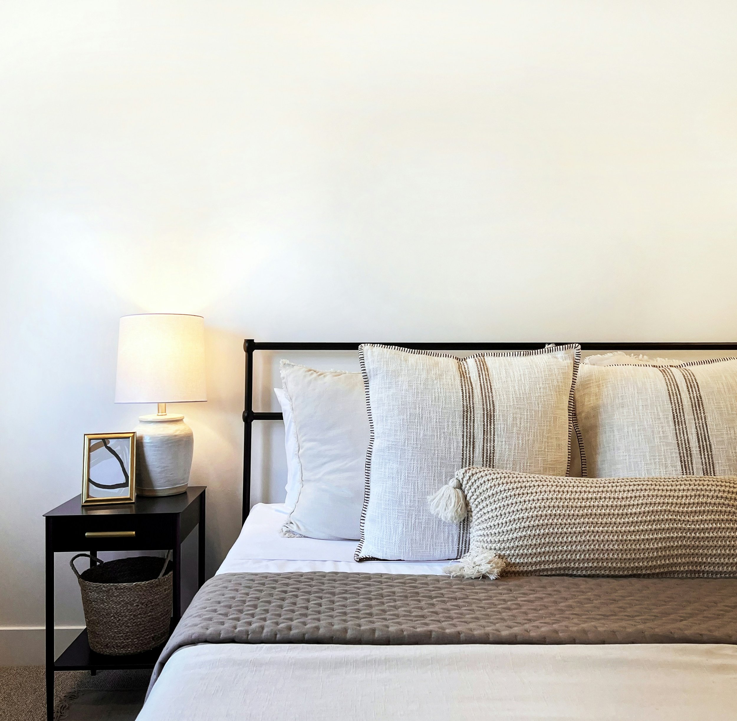 A bedroom with a bed, pillow, and blanket, a black nightstand with a framed picture, a white lamp, and a woven basket underneath.