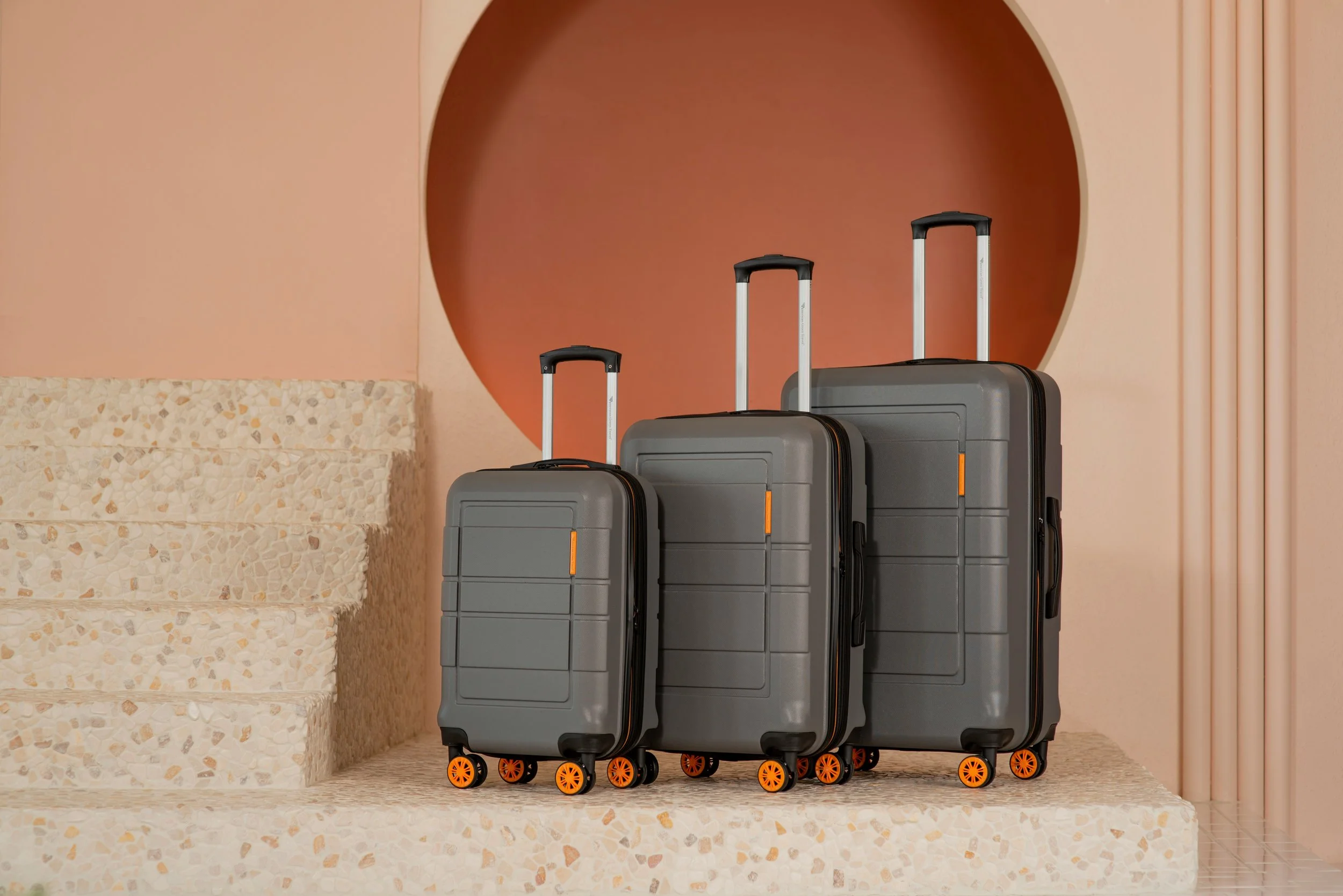 Five gray suitcases with orange wheels stacked in front of a pink wall with a large circular cutout and a beige speckled bench.