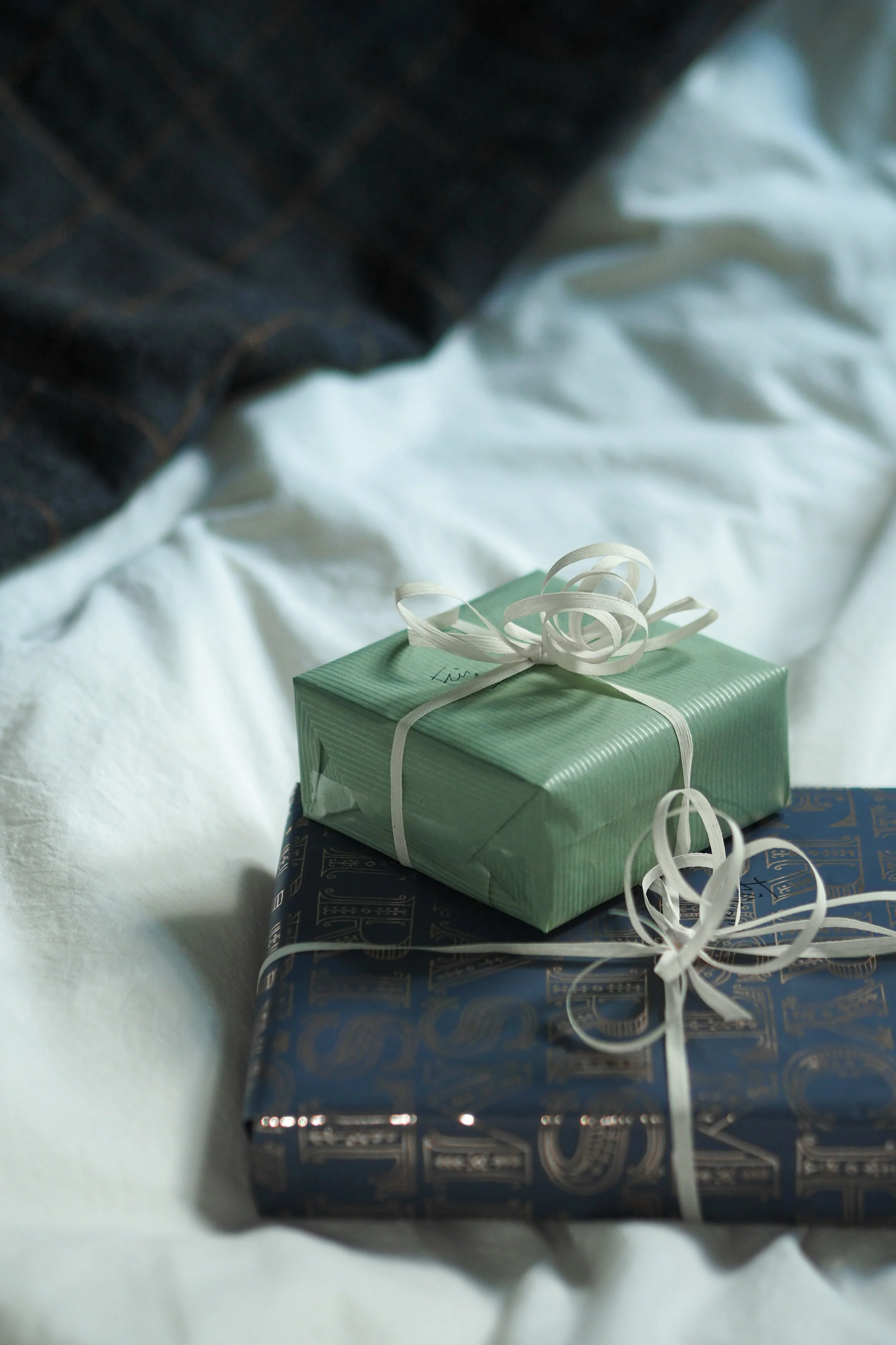 Three wrapped presents stacked on a white surface with ribbons and tags, an out of focus dark blue gift wrap at the bottom, a green gift with a white ribbon on top, and a larger dark blue gift with a gold pattern.