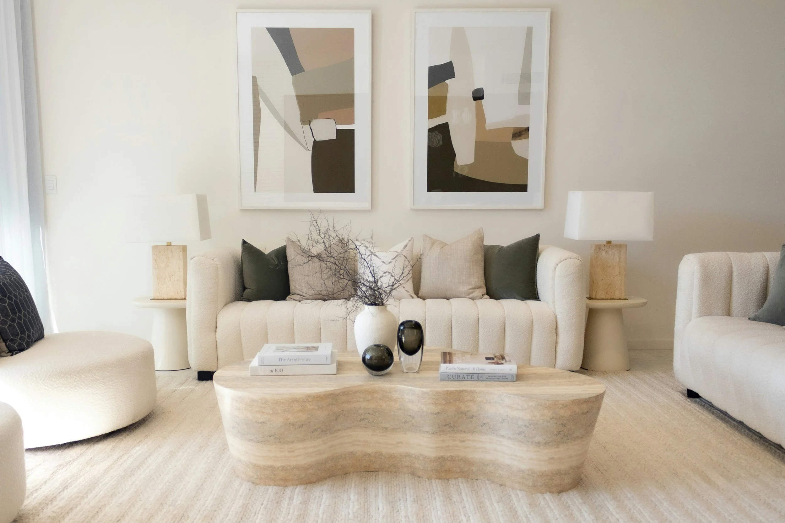 Living room with white sofa, black and beige pillows, two beige side tables with white lamps, two abstract framed artworks on the wall, a light-colored wooden coffee table with magazines and decorative objects, neutral color scheme with beige and white tones, and a textured rug.