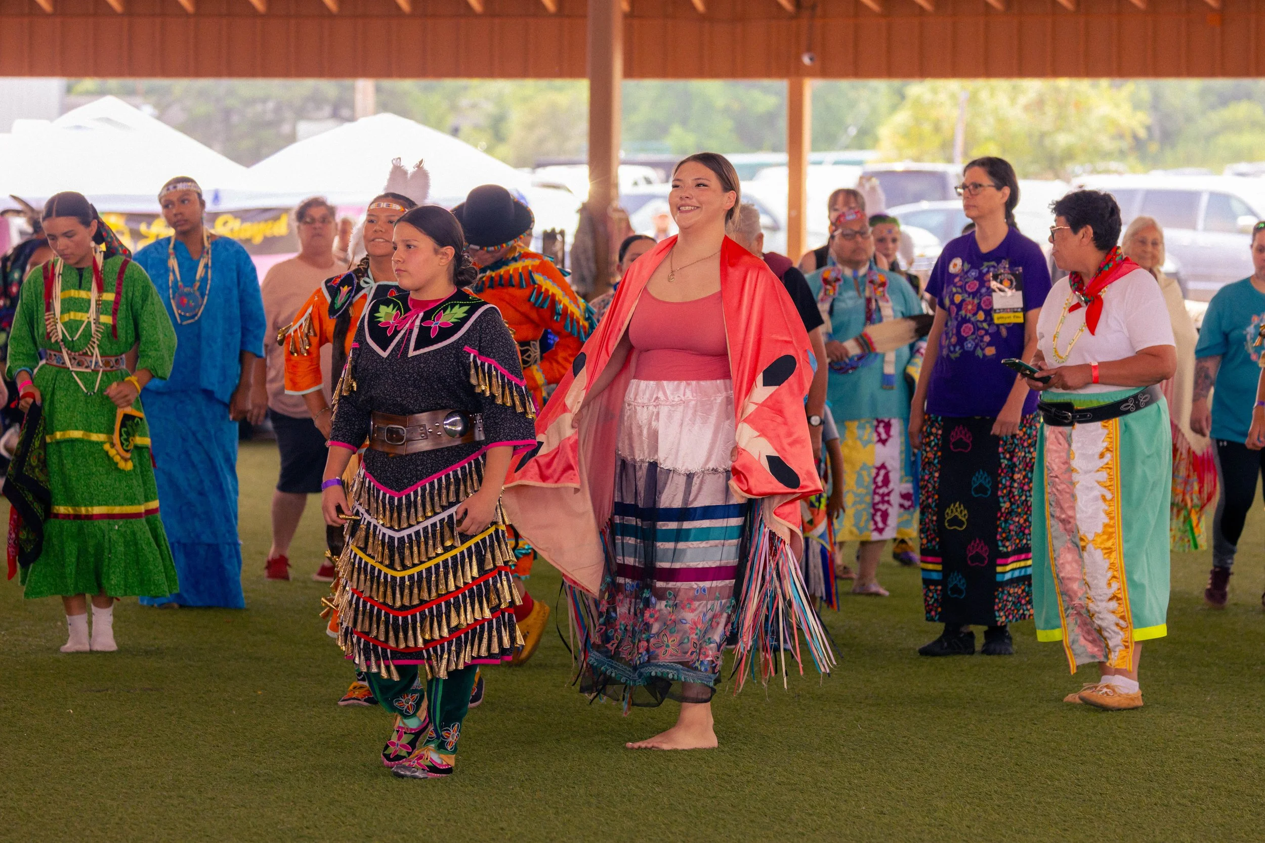 Group of Native American women and girls dressed in traditional regalia, dancing indoors at a cultural event.