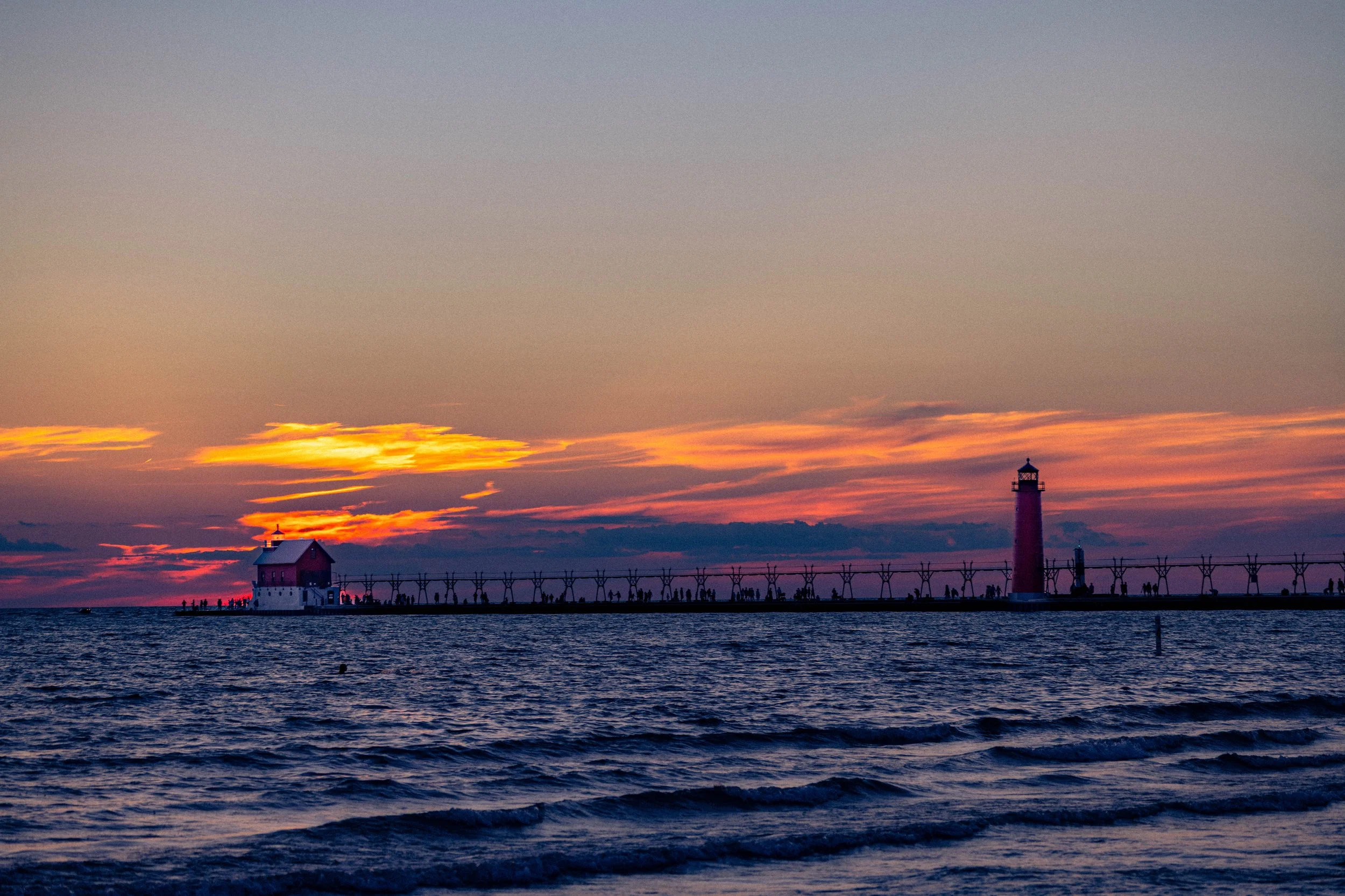A sunset over water with a lighthouse and a pier with people walking.