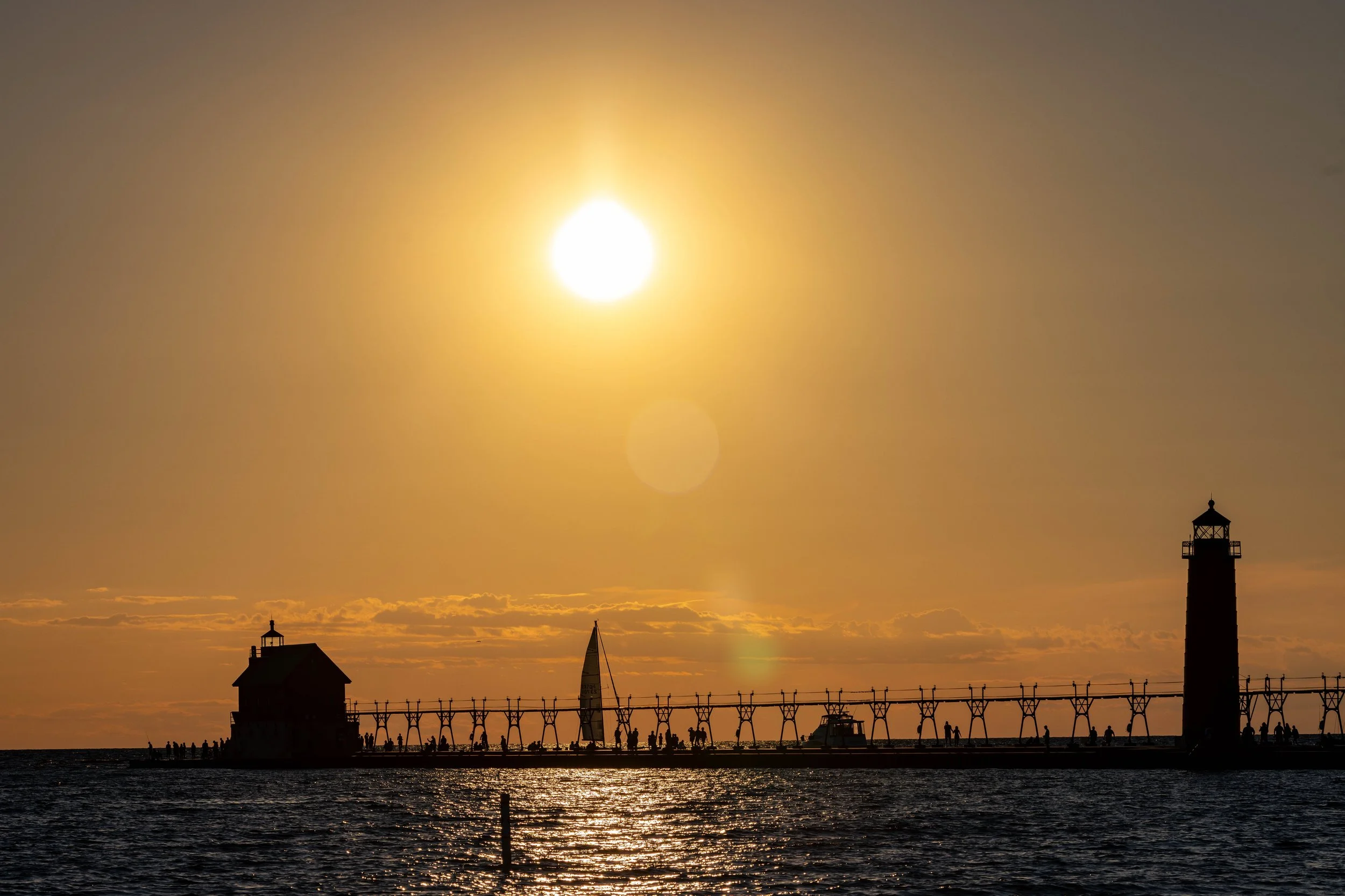 Sun setting over a pier with lighthouse and boats, silhouetted against the sky, with reflections on the water.