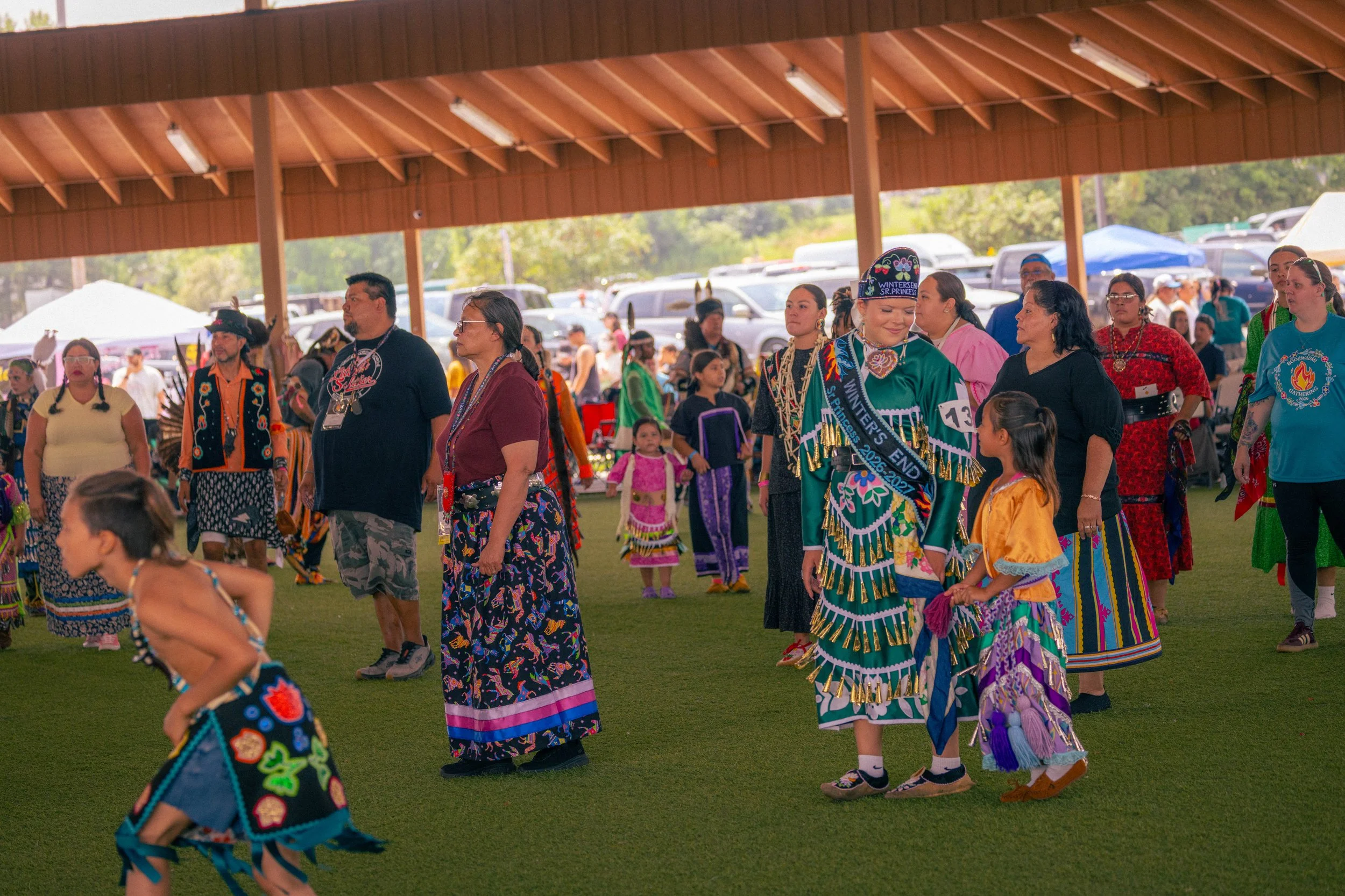 A group of people in traditional Native American clothing gathered under a covered outdoor pavilion, participating in a cultural event or dance.