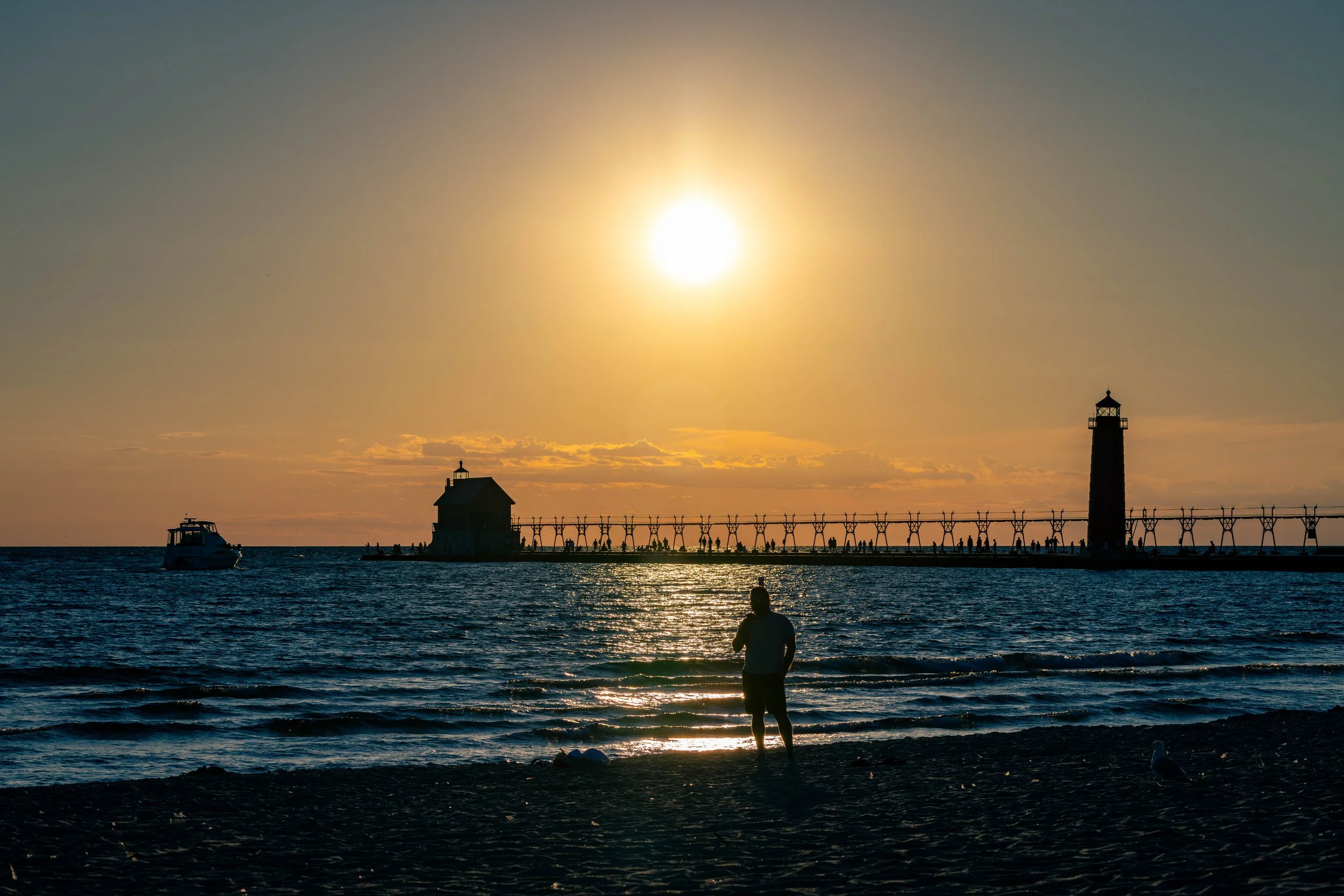 A sunset over the ocean with a lighthouse on a pier, boats, and a person in the water with seagulls.