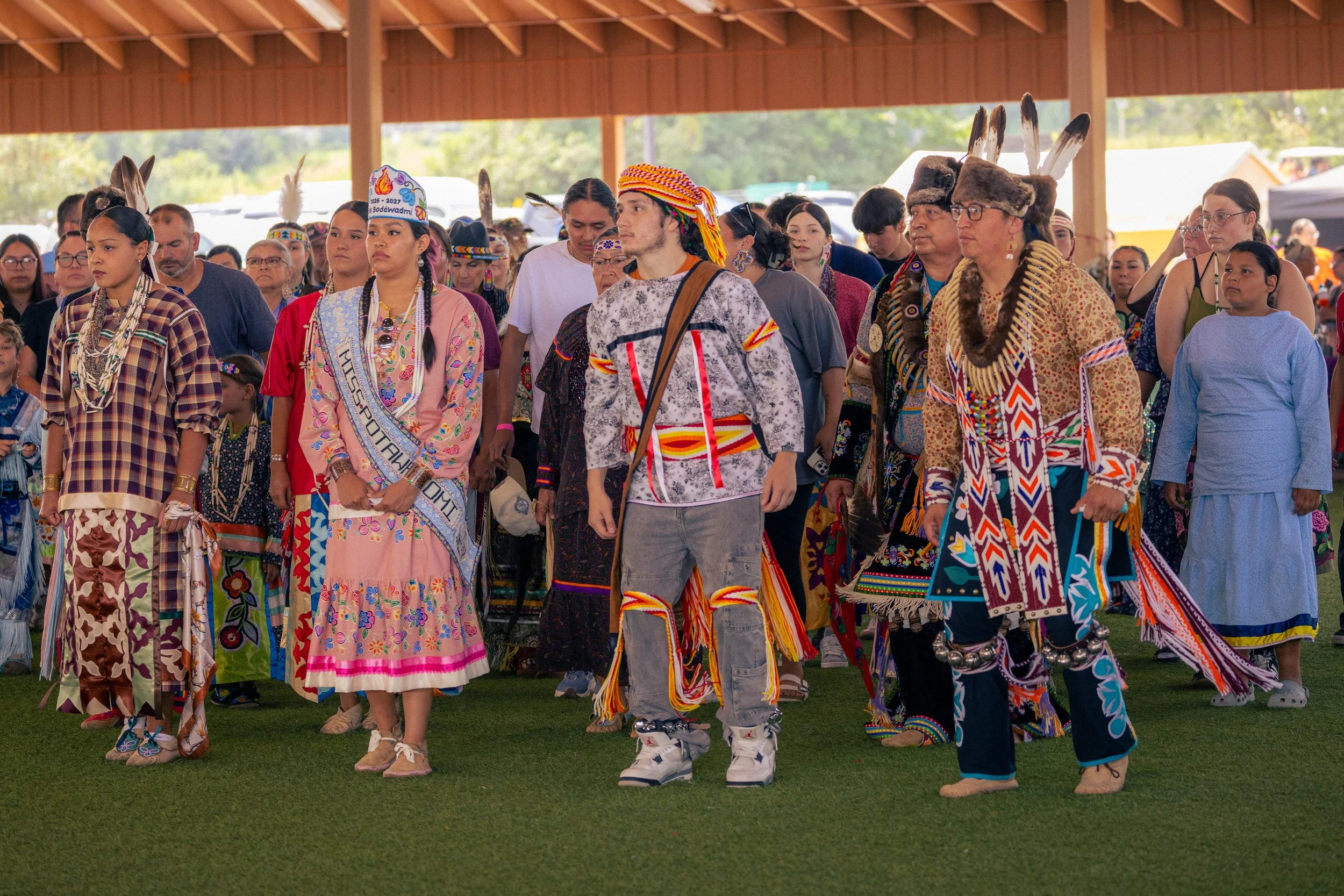 Group of Indigenous people dressed in traditional clothing, dancing or parading indoors at a cultural event.