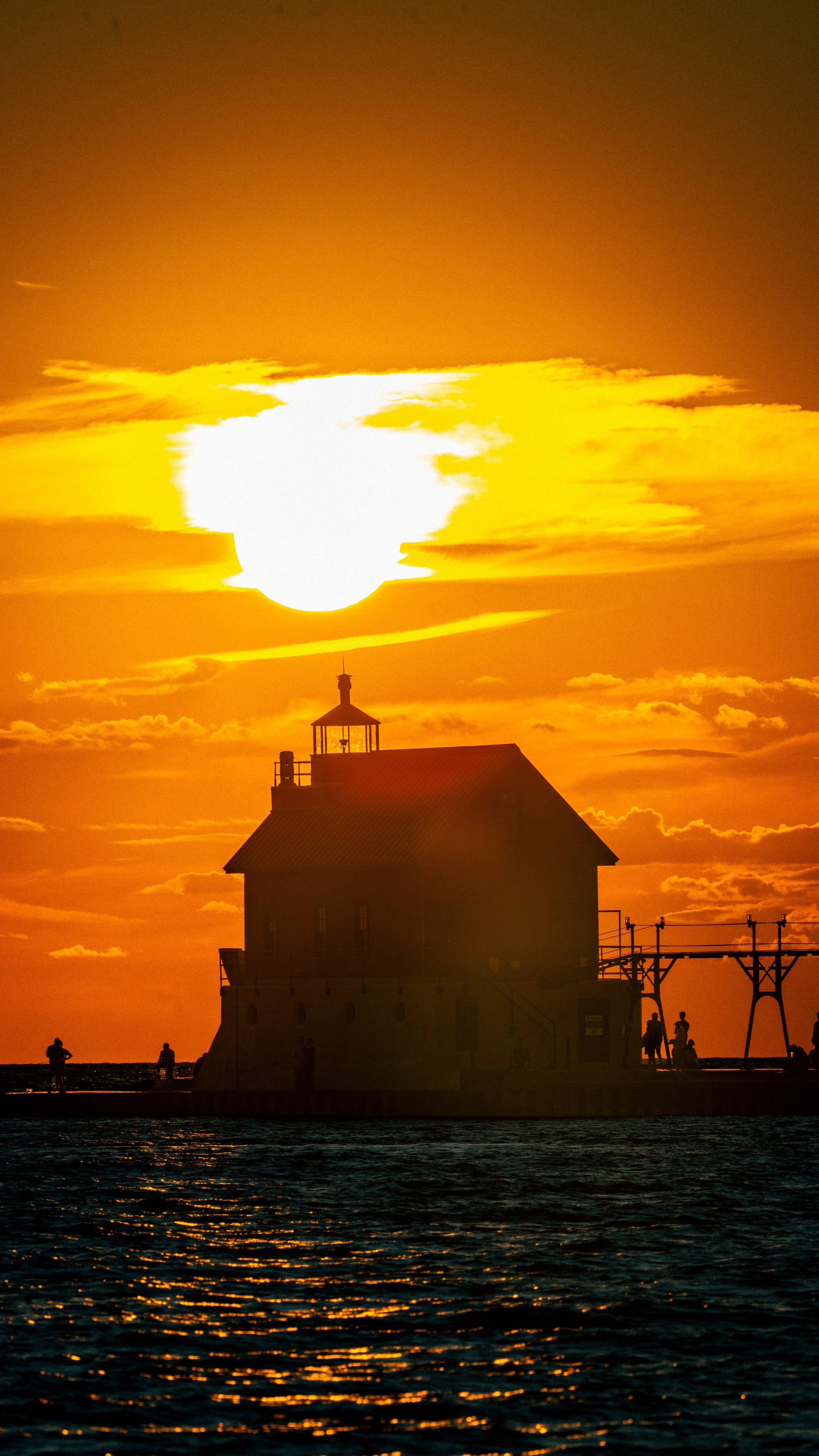 Sunset over a lighthouse on water with a small silhouette of people on a dock, vibrant orange sky with clouds.
