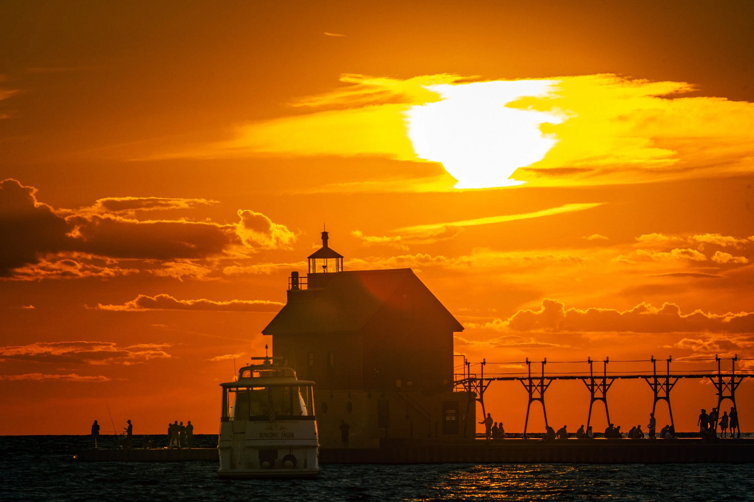 Sunset over a harbor with silhouettes of people, a boat, a lighthouse building, and a bridge, with the sky in vibrant orange and yellow hues.