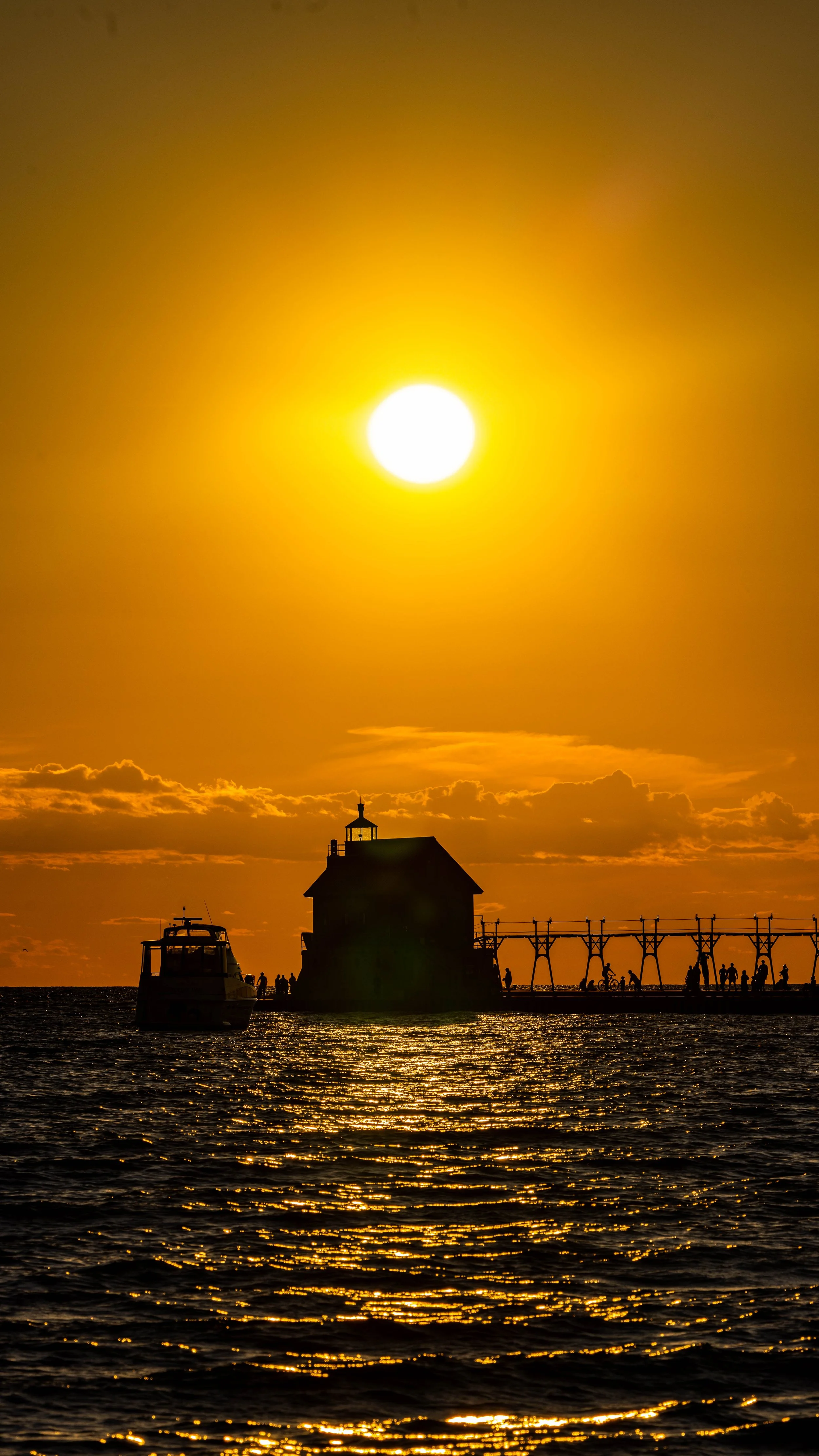 Sunset over a harbor with boats, a lighthouse, and a pier with people walking and cycling.