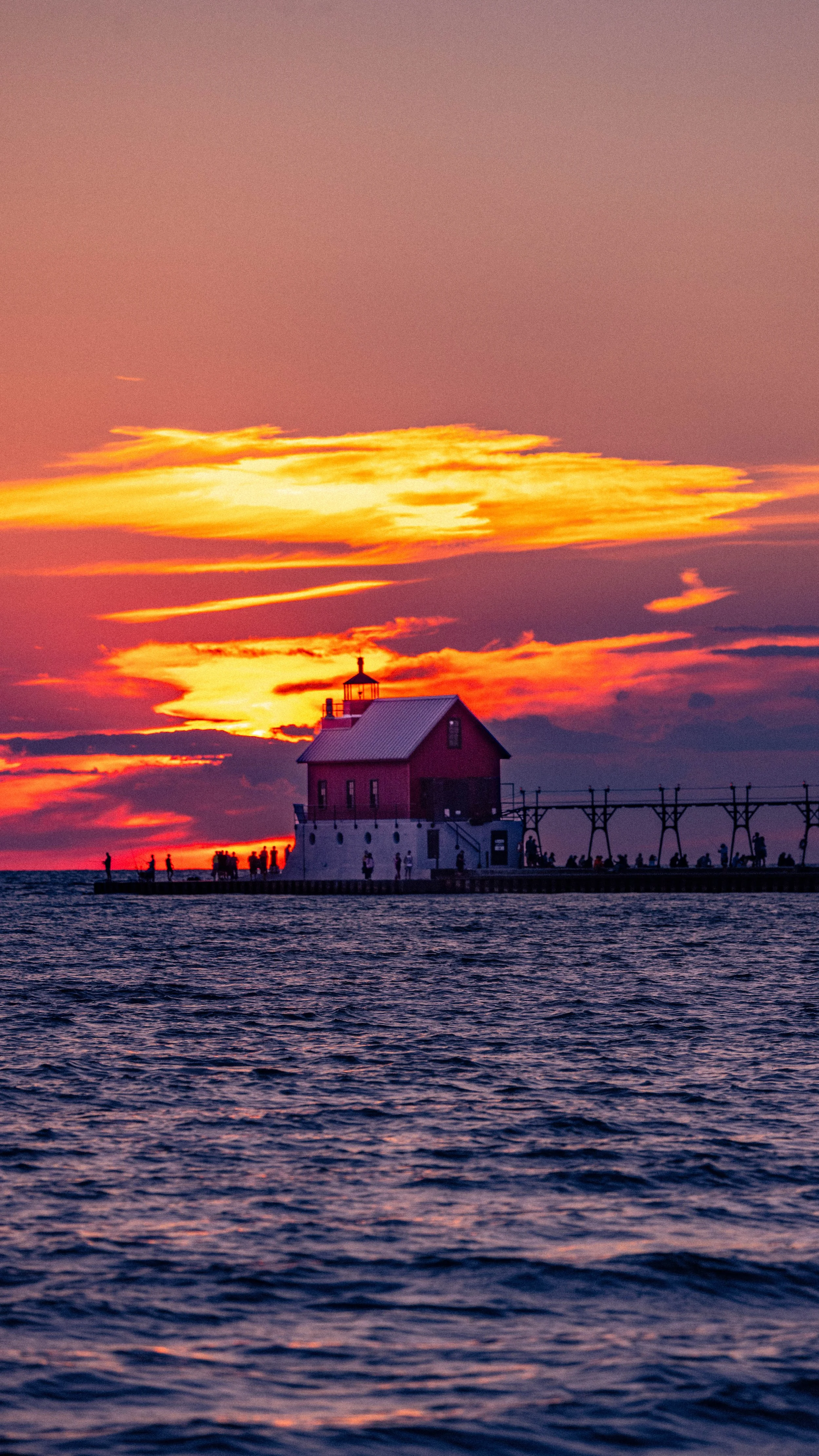 A lighthouse on a pier during sunset, with a colorful sky and silhouettes of people near the lighthouse.