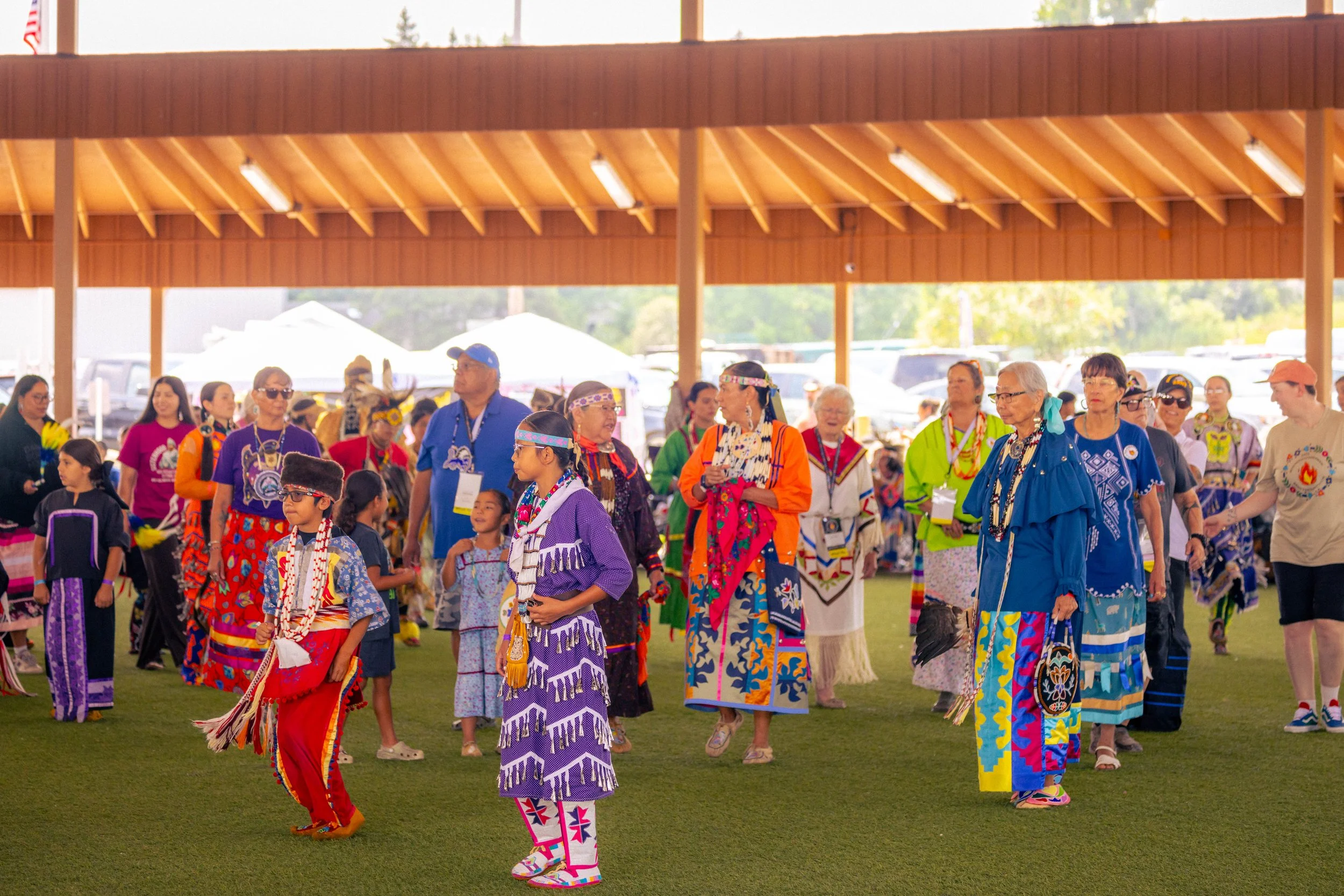 Group of Indigenous people dressed in colorful traditional clothing, standing and interacting at a cultural event under a wooden pavilion.