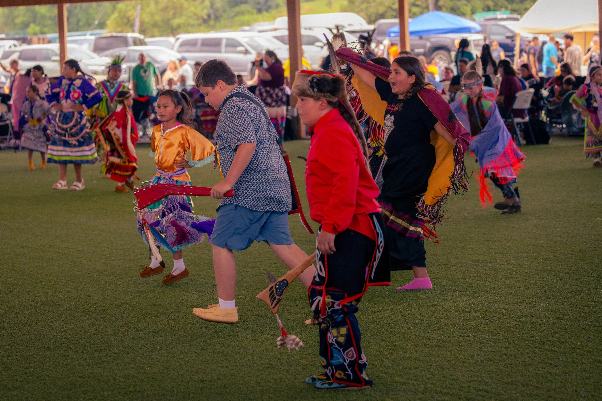 Children dressed in traditional Native American clothing participating in a cultural dance at an outdoor event with a crowd and parked cars in the background.