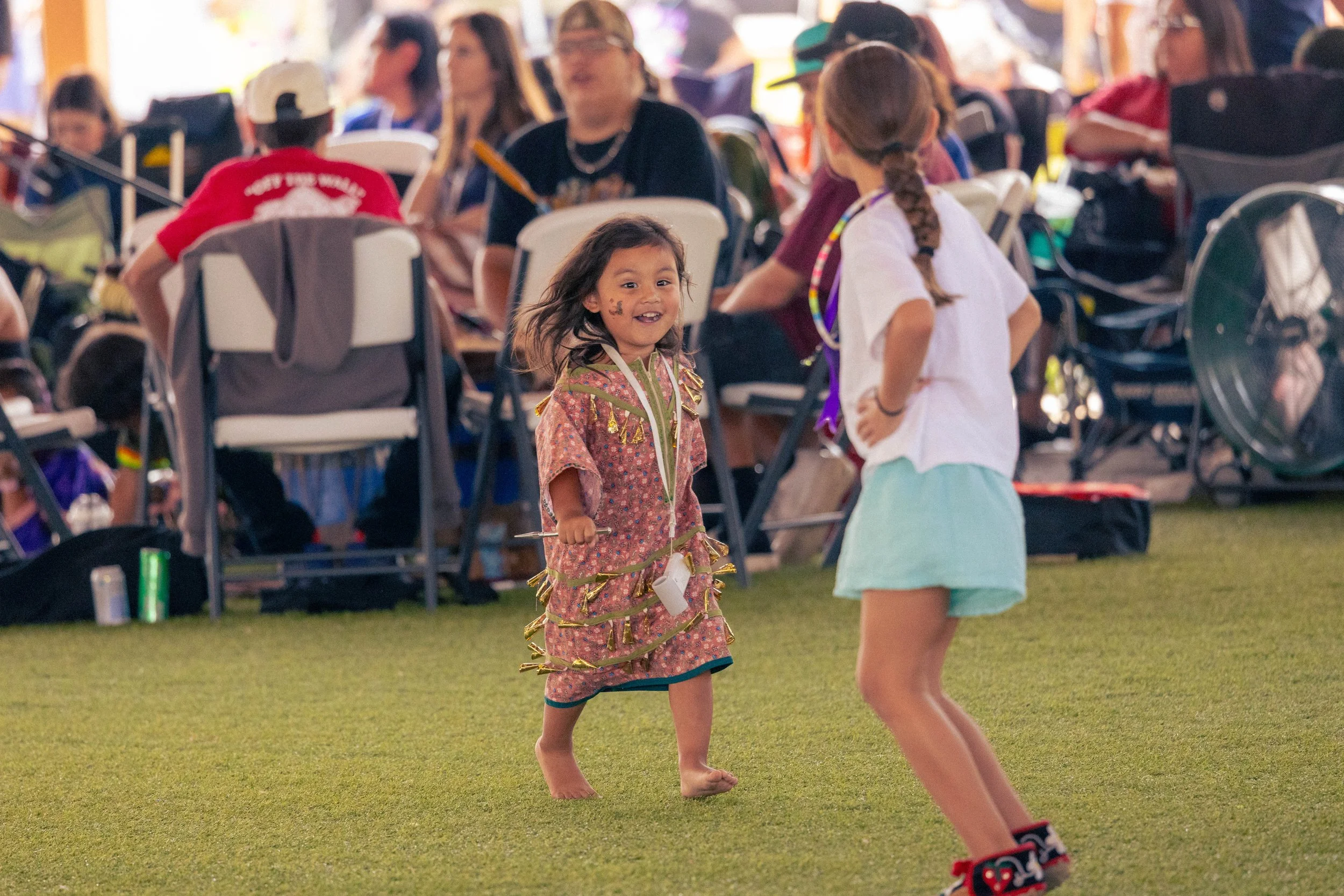Young girl smiling and playing with streamers at a crowded outdoor event, with people sitting and standing in the background.