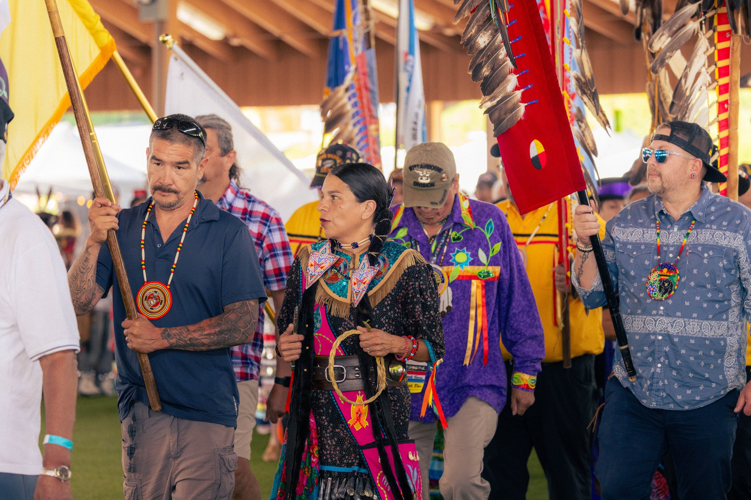 A group of people participating in a Native American or Indigenous cultural parade or event. They are dressed in traditional attire, with some holding flags, and standing under a covered structure.