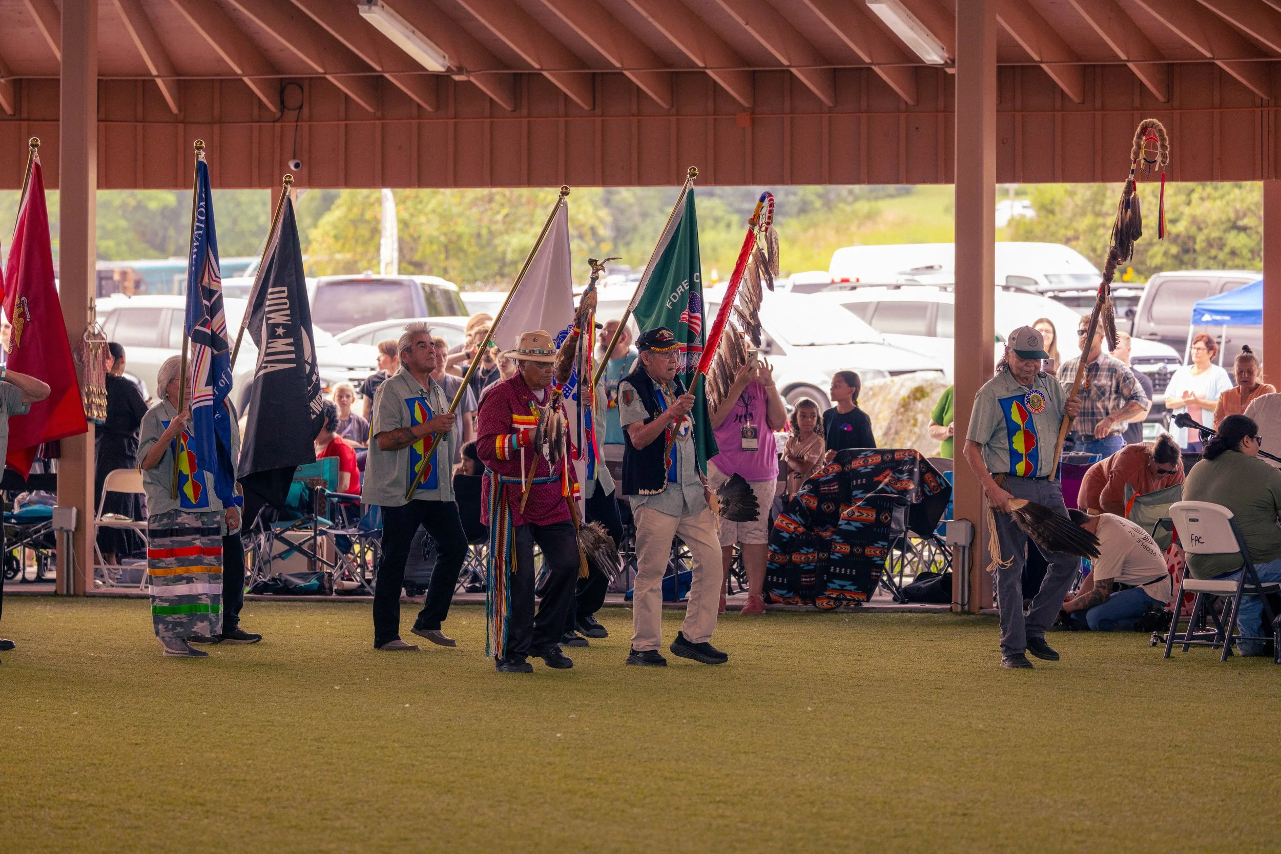 A group of Native Americans in traditional attire carrying flags in a cultural event under a pavilion at an outdoor venue.
