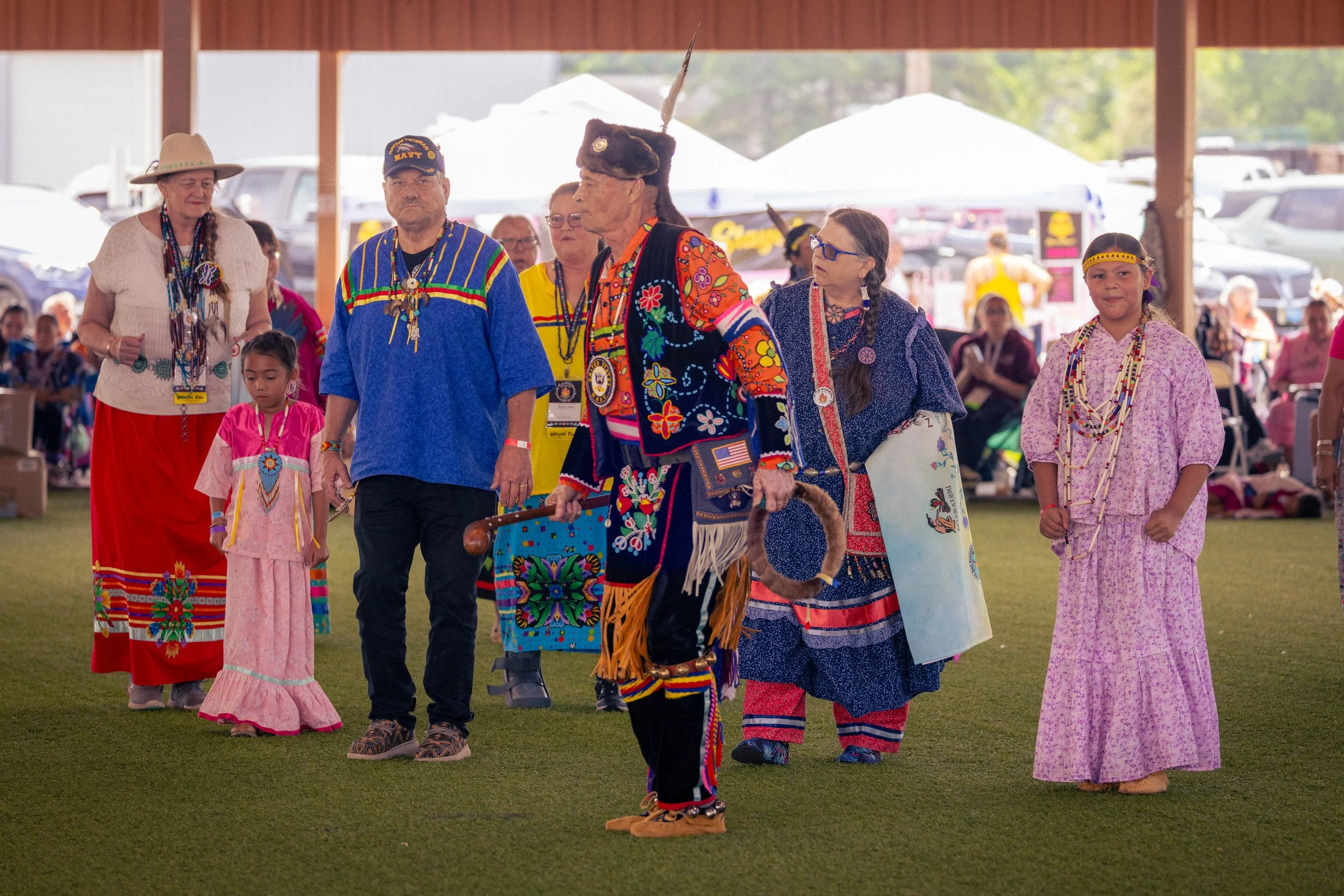 Group of Indigenous people dressed in traditional clothing, walking in a line at an outdoor cultural event under a pavilion with parked cars and tents in the background.