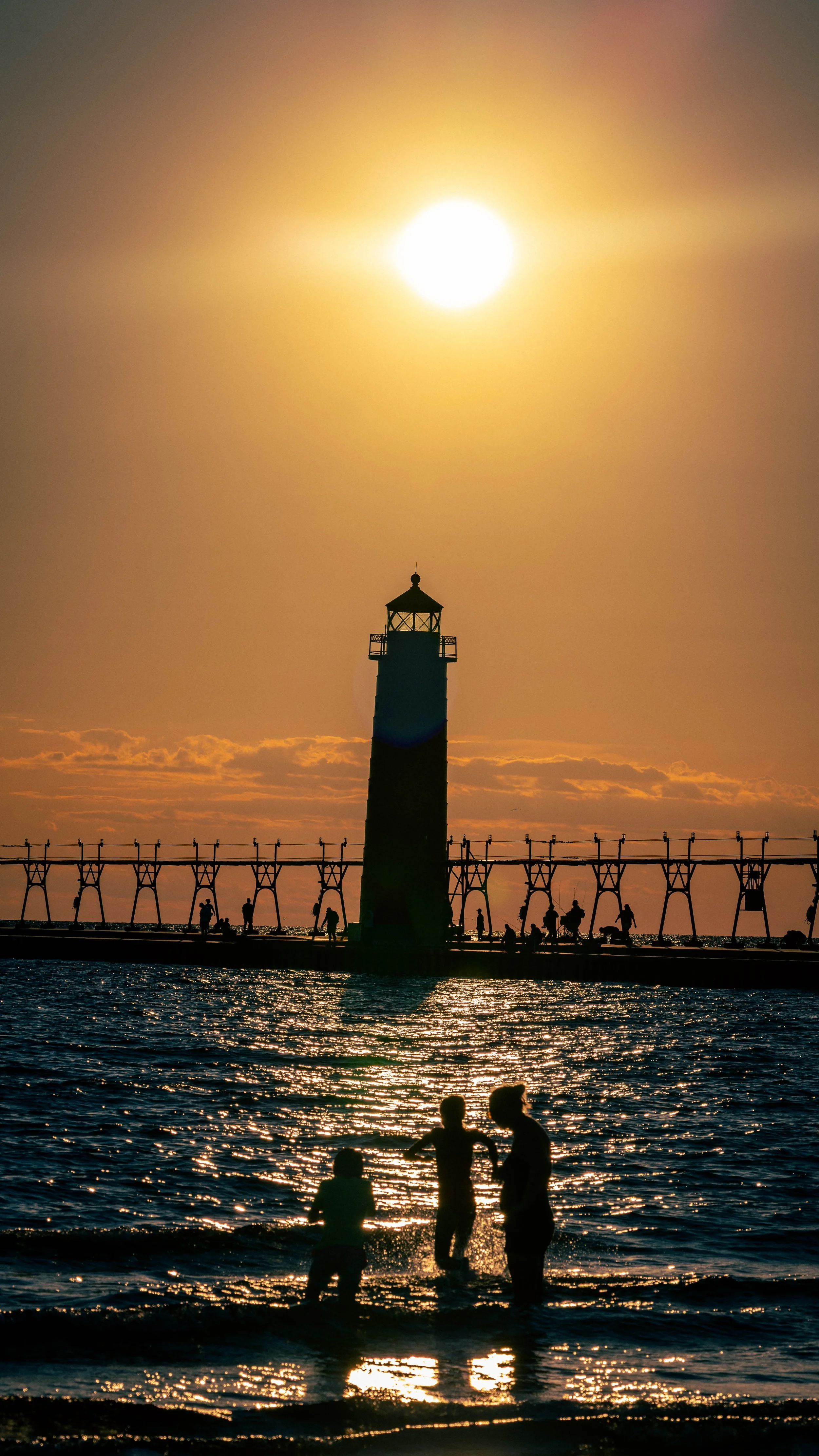 Silhouettes of children playing by the water with a lighthouse and a pier in the background, during sunset.