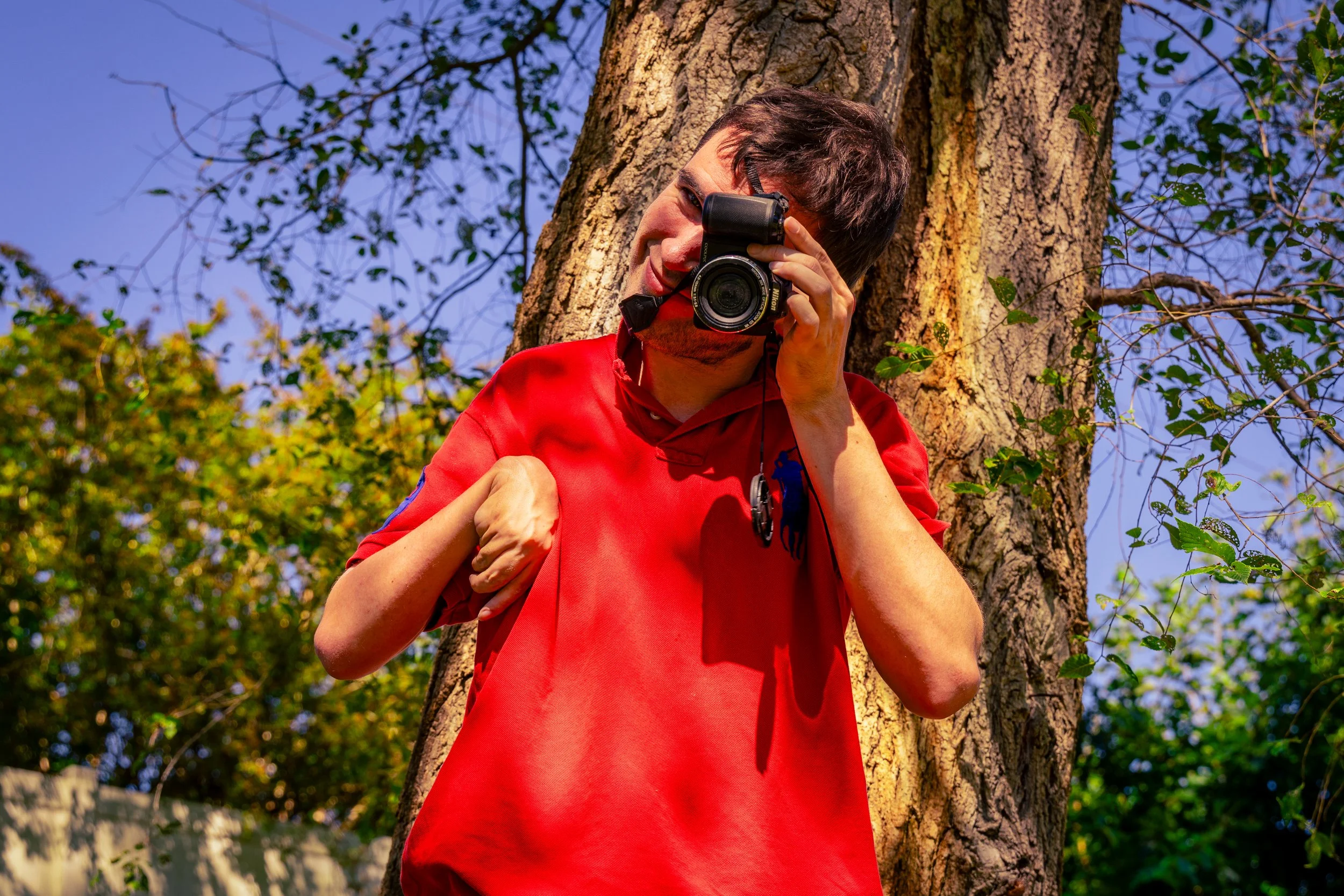 A man wearing a red shirt is taking a photograph with a camera outdoors near a tree. The background features a blue sky and green foliage.