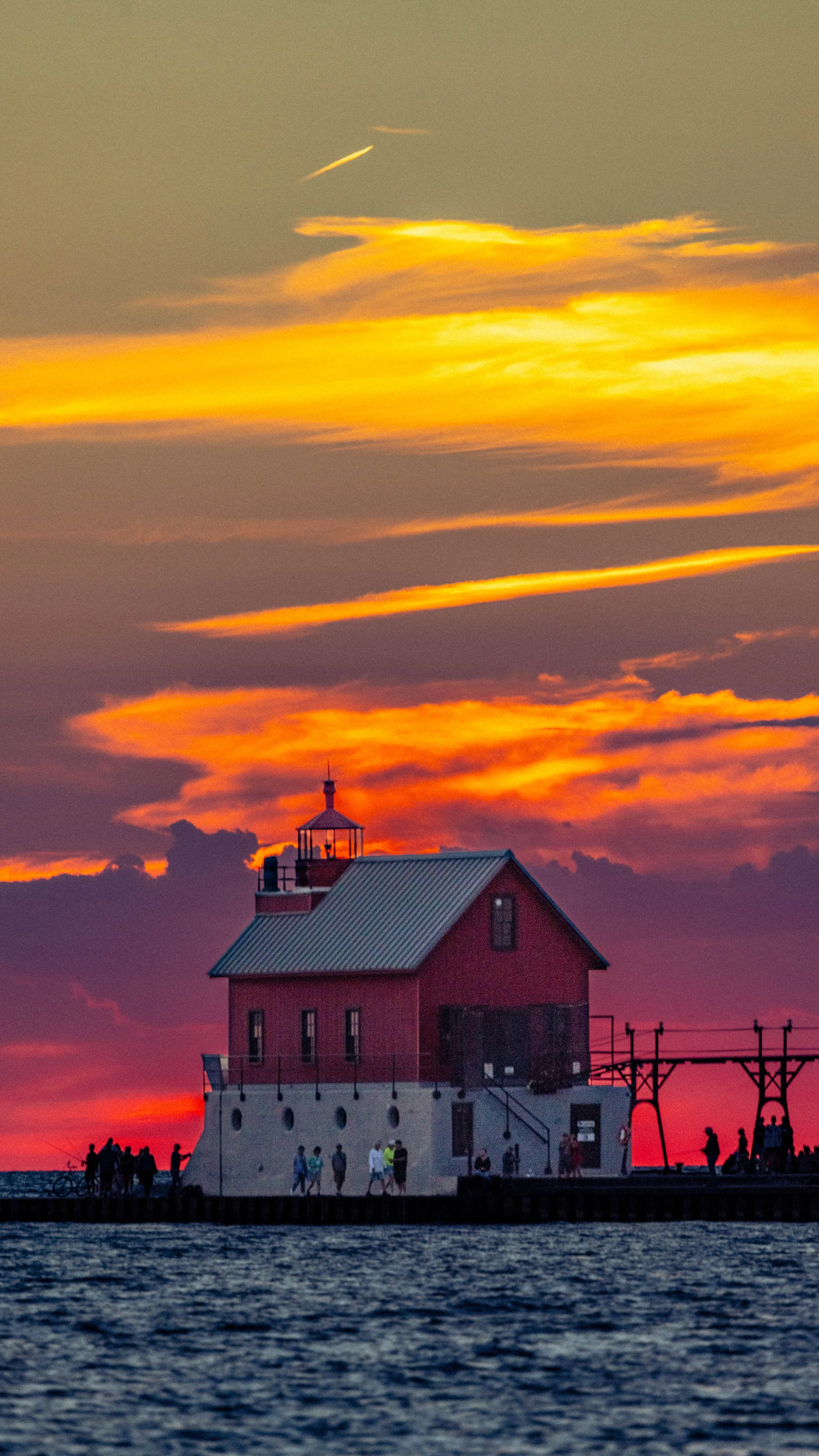 A lighthouse on a pier with a red building, people walking and sitting nearby, under a colorful sunset sky with orange and yellow clouds.